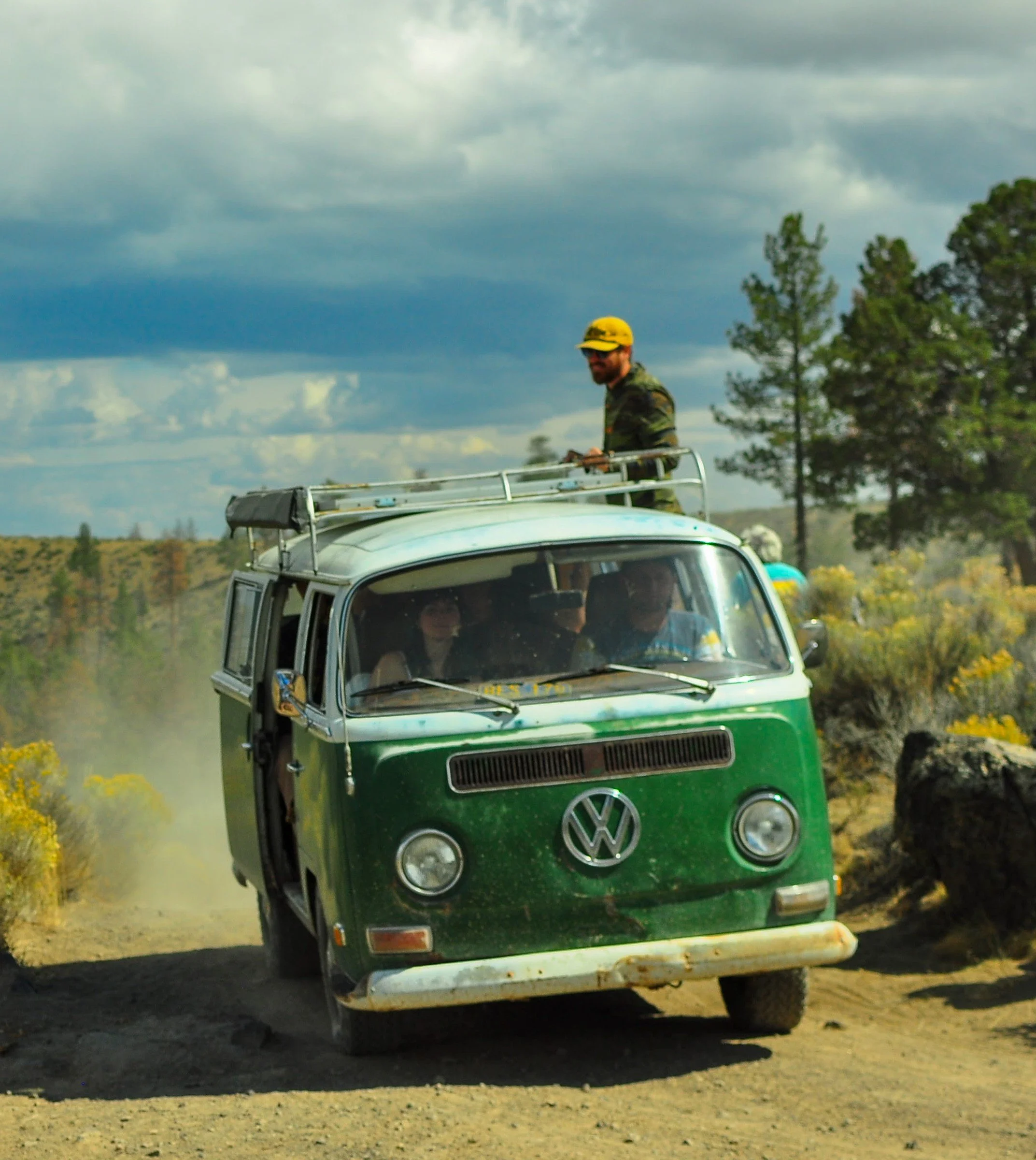 A vintage green Volkswagen van driving on a dirt road in a rural area with trees and bushes, under a cloudy sky, with a person standing on the roof wearing a yellow hat. Oregon, Descend on Bend 2023.