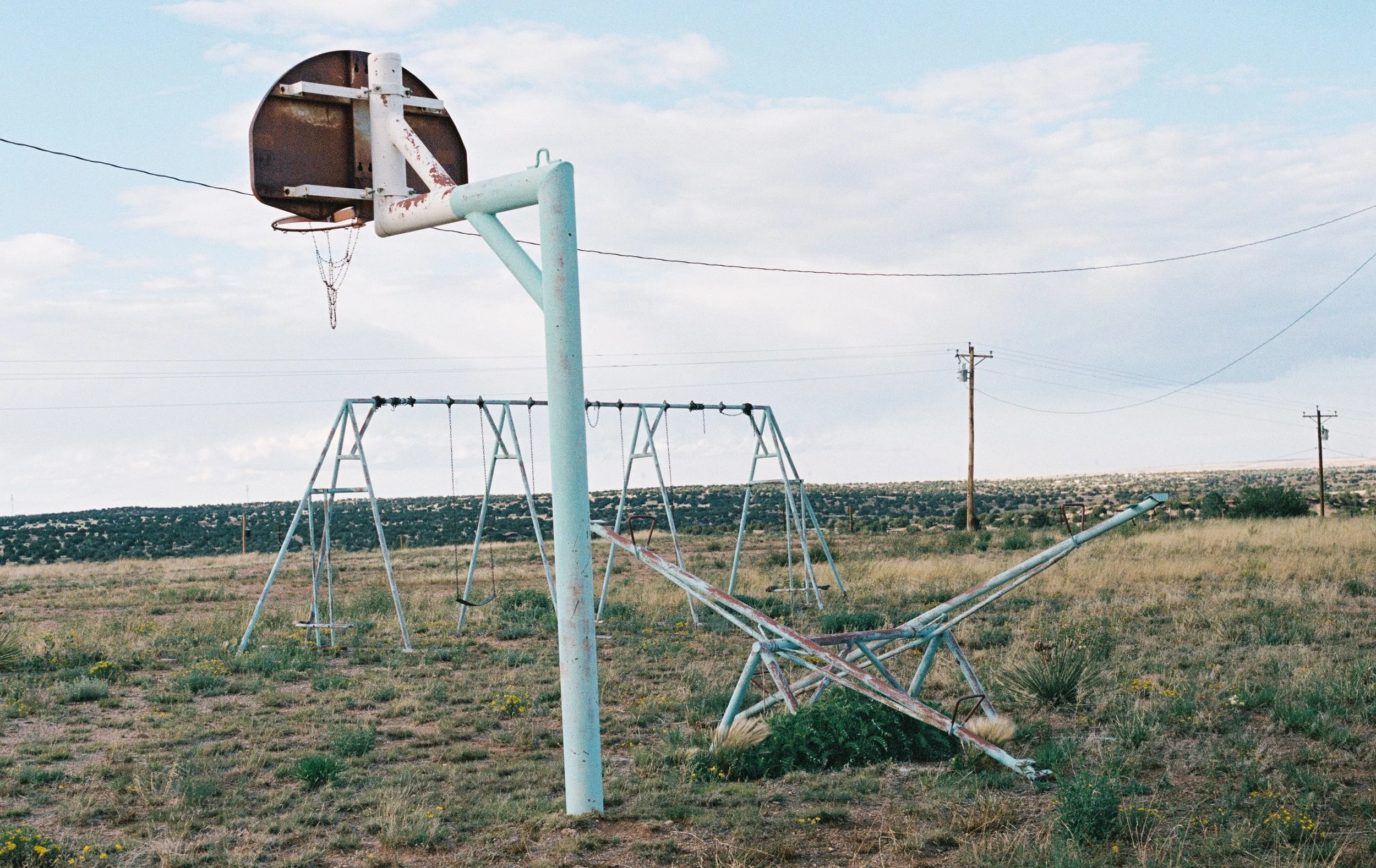 An abandoned playground with a rusted swing set, a tilted slide, and a broken basketball hoop in a grassy field under a cloudy sky. Route 66