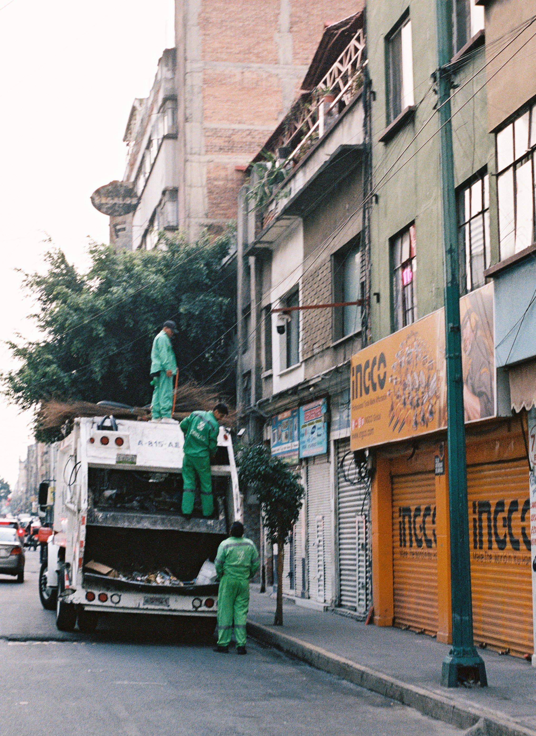 Workers in green uniforms collecting trash from a garbage truck in Mexico City, with closed stores and apartment buildings in the background.