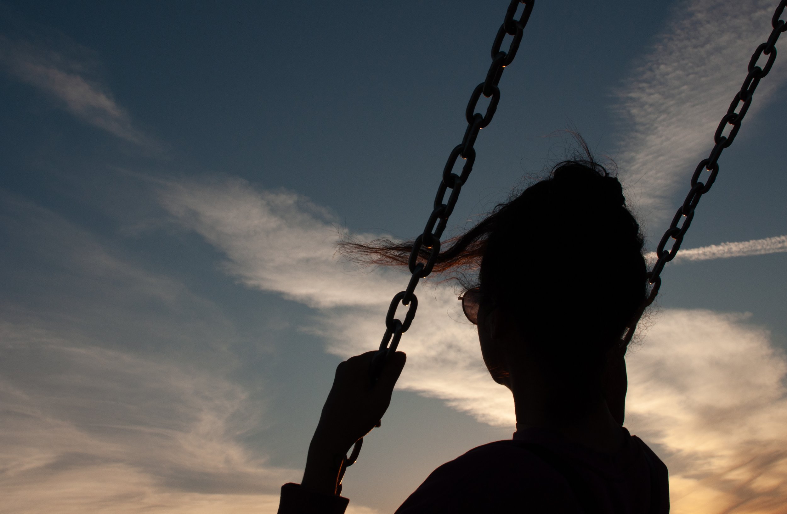 Silhouette of a person sitting on a swing against a sunset sky with clouds and contrails.