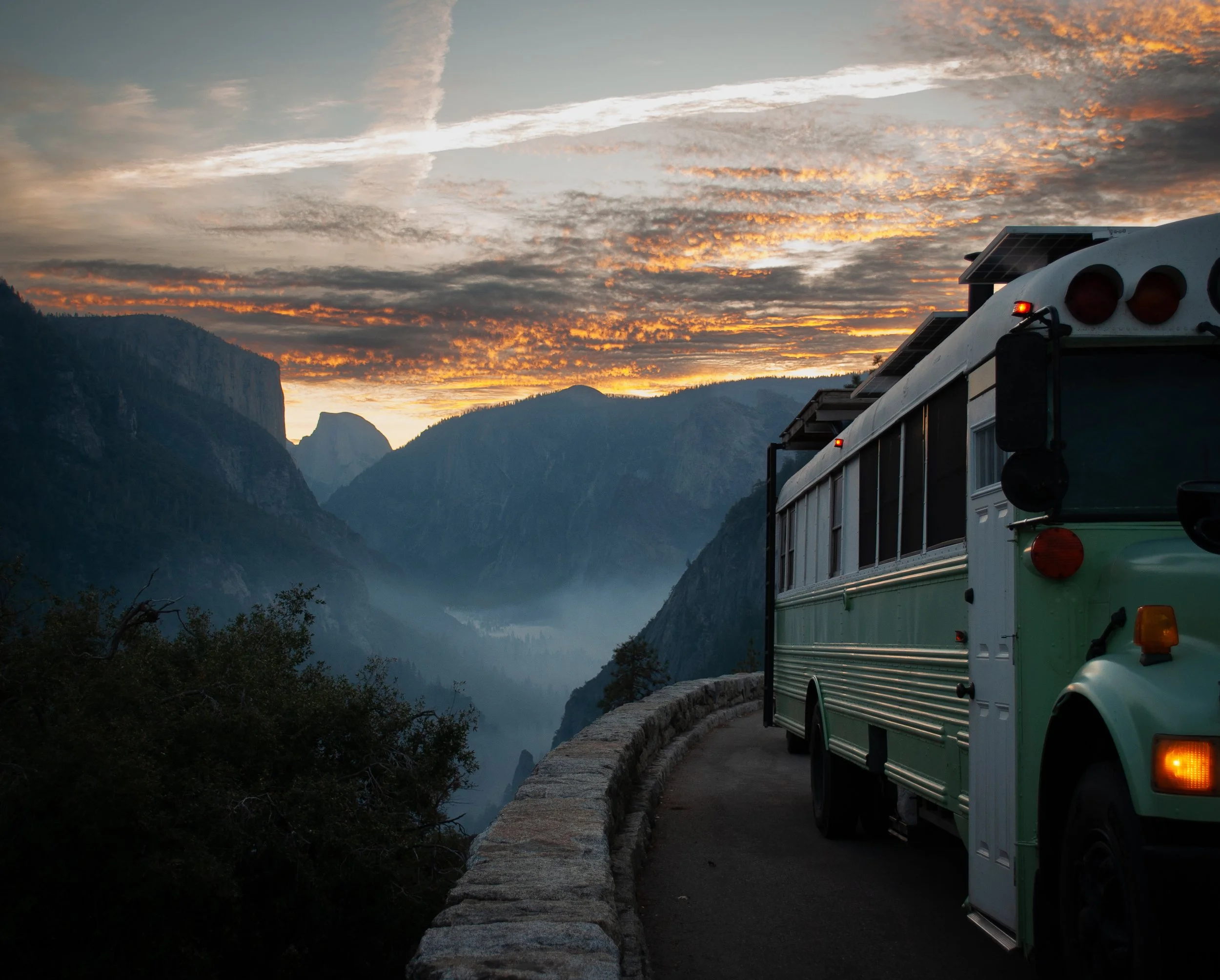 A skoolie parked along a mountain road at sunrise, overlooking Yosemite Valley with the famous geological landmark, "Half Dome" seen in the background