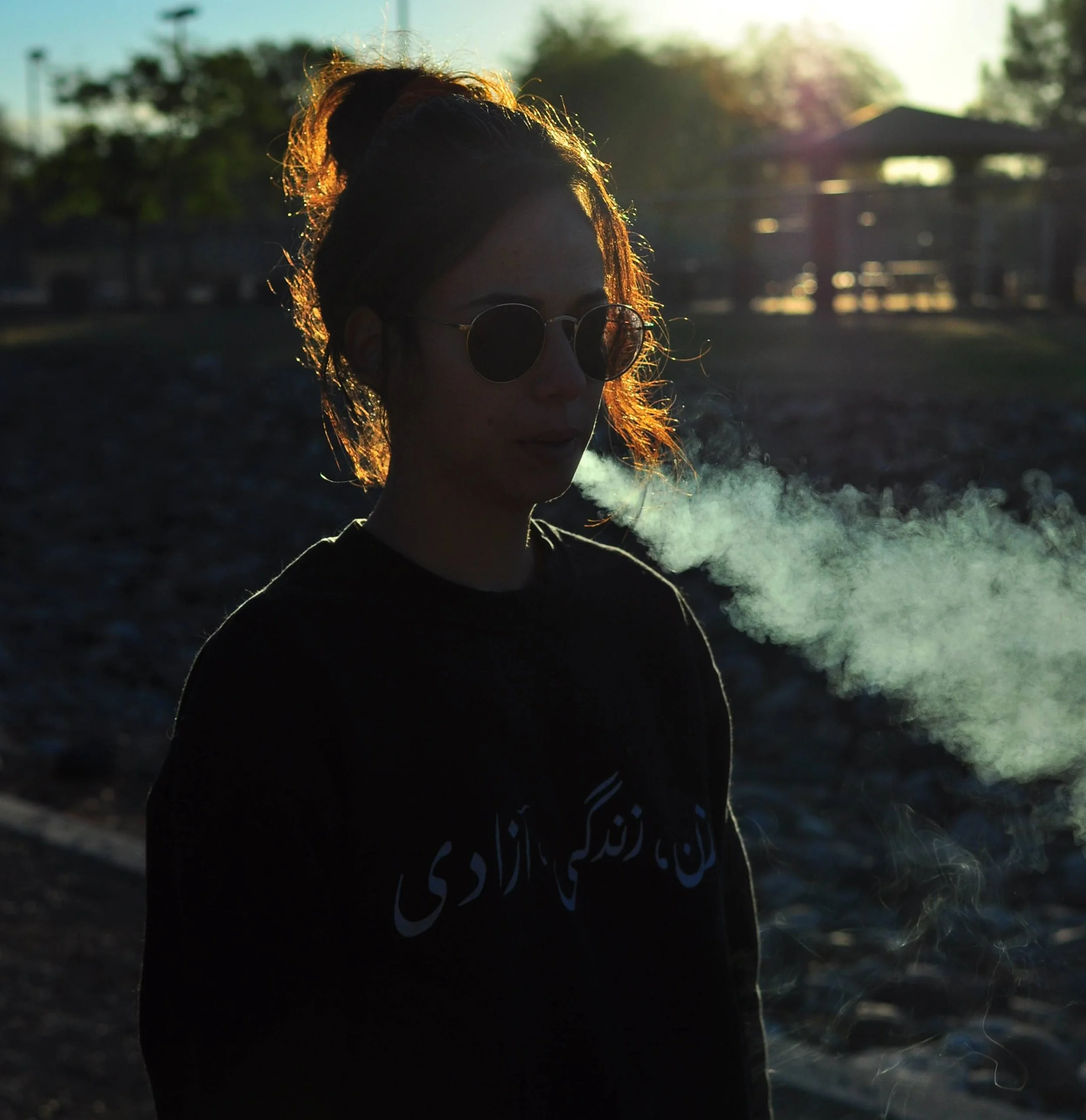 Silhouette of a young woman wearing sunglasses blowing smoke from her mouth, standing outdoors during sunset or sunrise. Arabic
