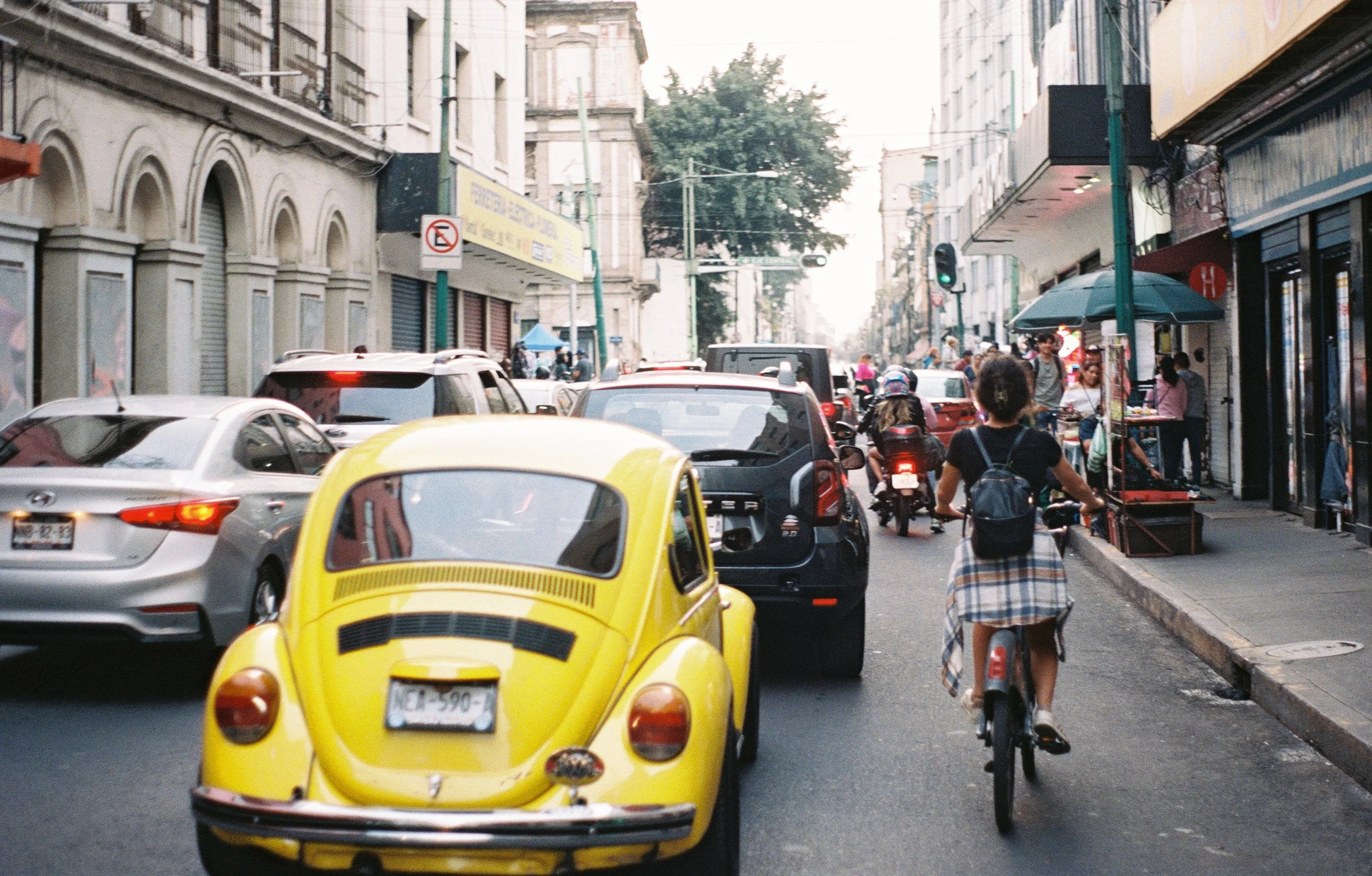Mexico City street scene with cars, a yellow vintage Volkswagen Beetle, a cyclist, pedestrians, street vendors, and urban buildings.