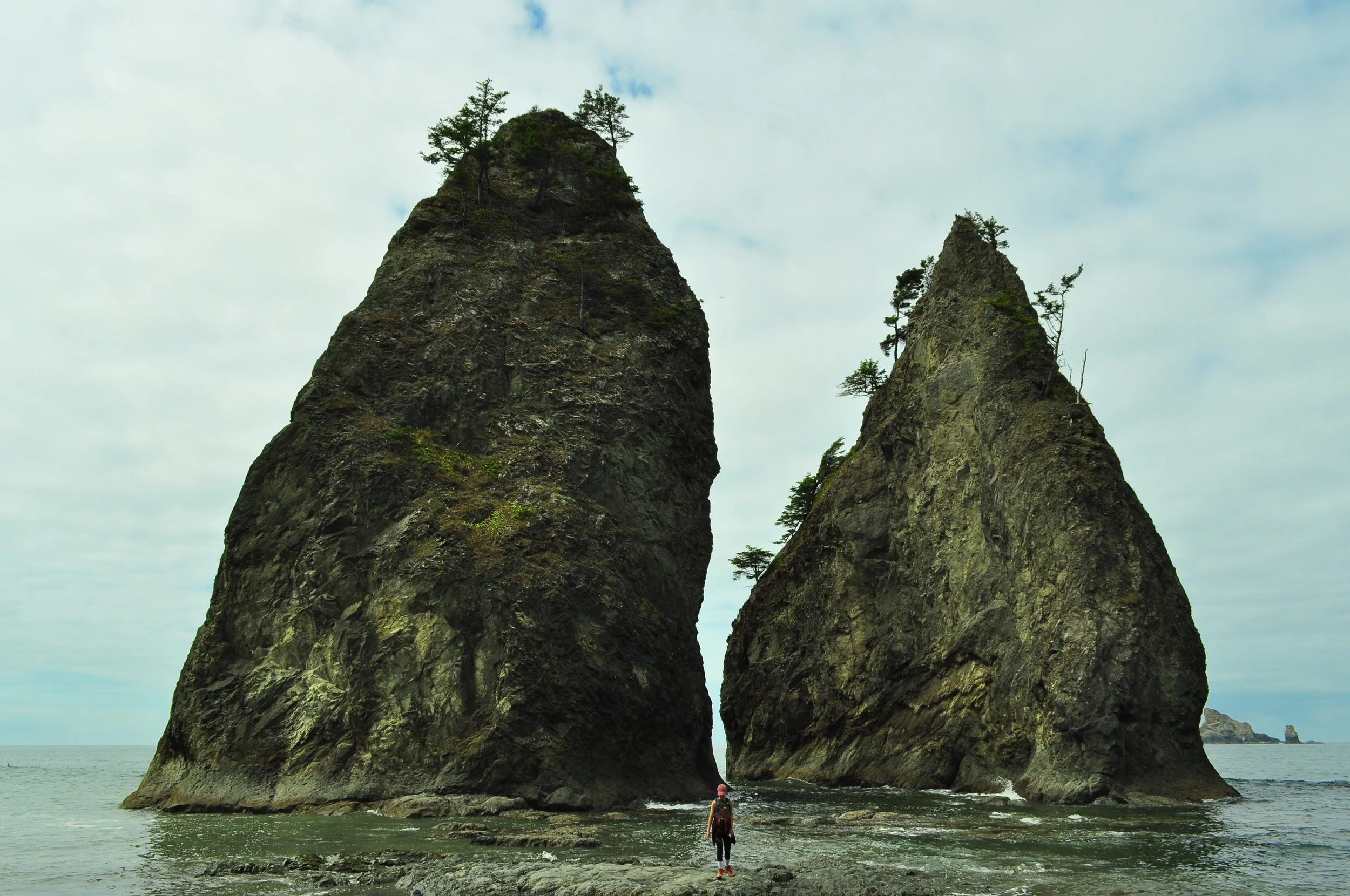 A person standing between Split Rock on a rocky shoreline with an ocean and cloudy sky in the background.