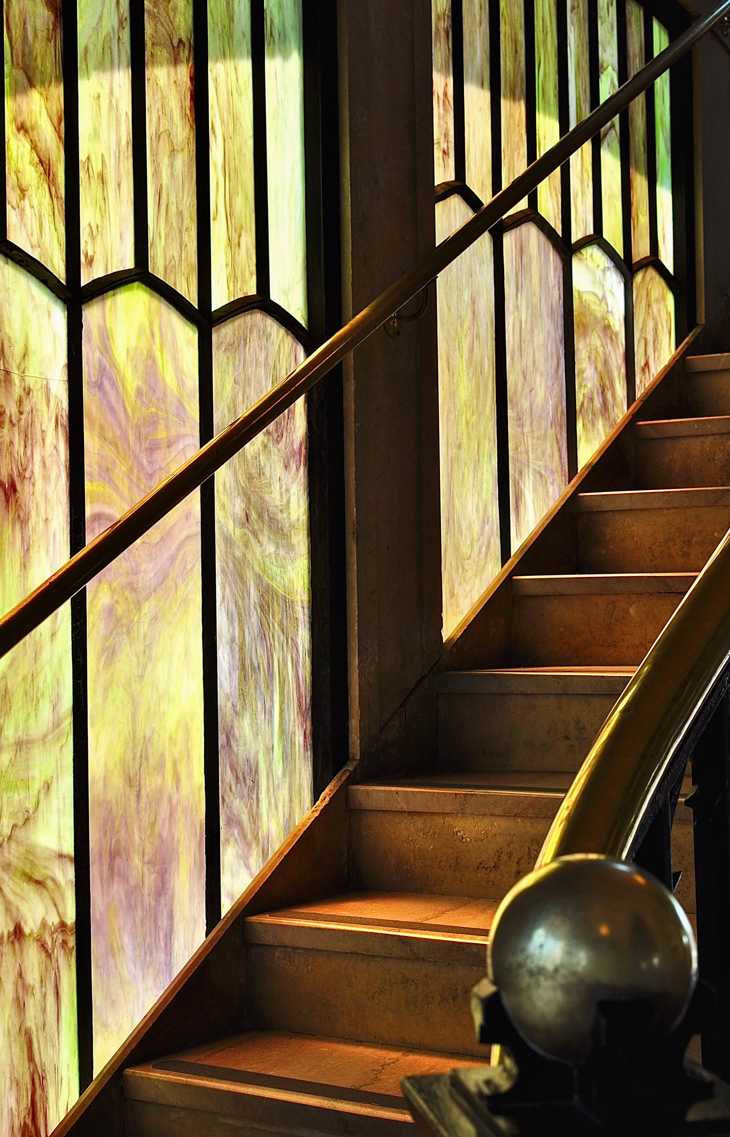 A staircase with a brass handrail beside a wall of colorful, abstract stained glass panels.