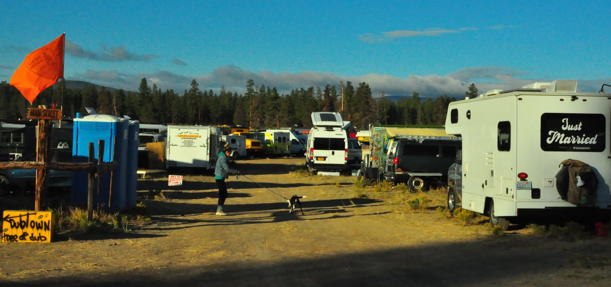 A woman walking a dog in a park with numerous RVs and camper vans, some with signs like 'Just Married', and a sign that reads 'DUBTOWN land of dub' Descend on Bend 2023 in Oregon with trees and mountains in the background.