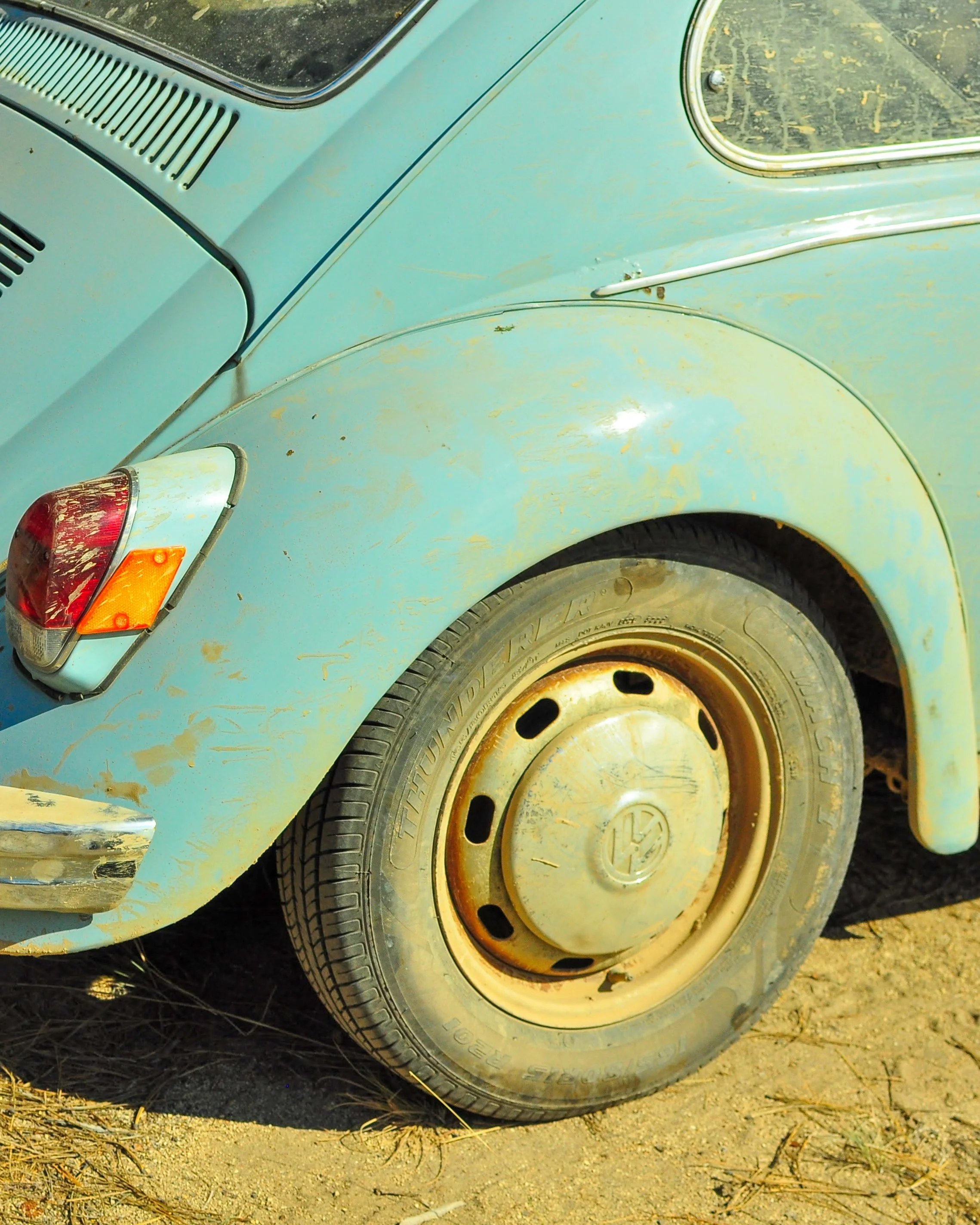 Close-up of the rear section of an old, weathered light blue Volkswagen Beetle, showing the rusty hubcap on the rear tire, part of the rear taillight, and the weathered body with rust spots and faded paint. Descend on Bend 2023.