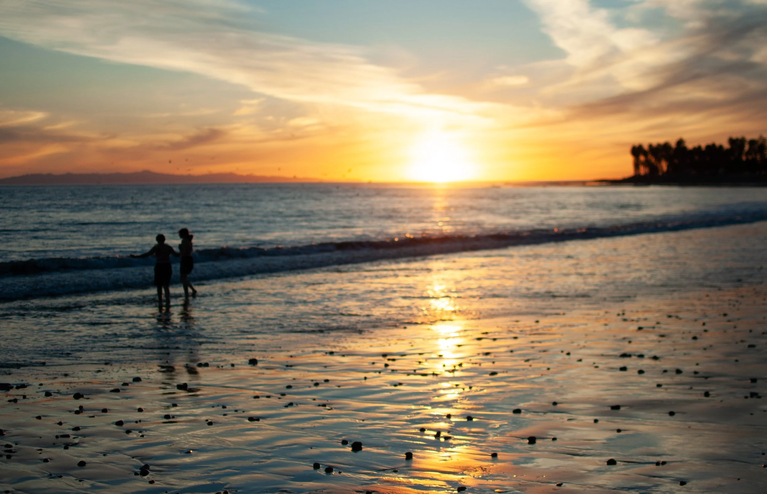 Two children playing in the shallow water at sunset on a beach, with a line of trees on the distant shoreline and a colorful sky with clouds. Ventura, California