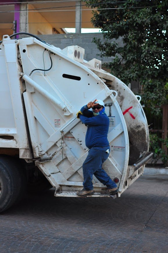 A man in a blue jacket and gray pants is standing on the open back of a garbage truck, saluting or shielding his eyes from the sun with his right hand, while wearing sunglasses and a white cap. The garbage truck is white and slightly dirty, parked on