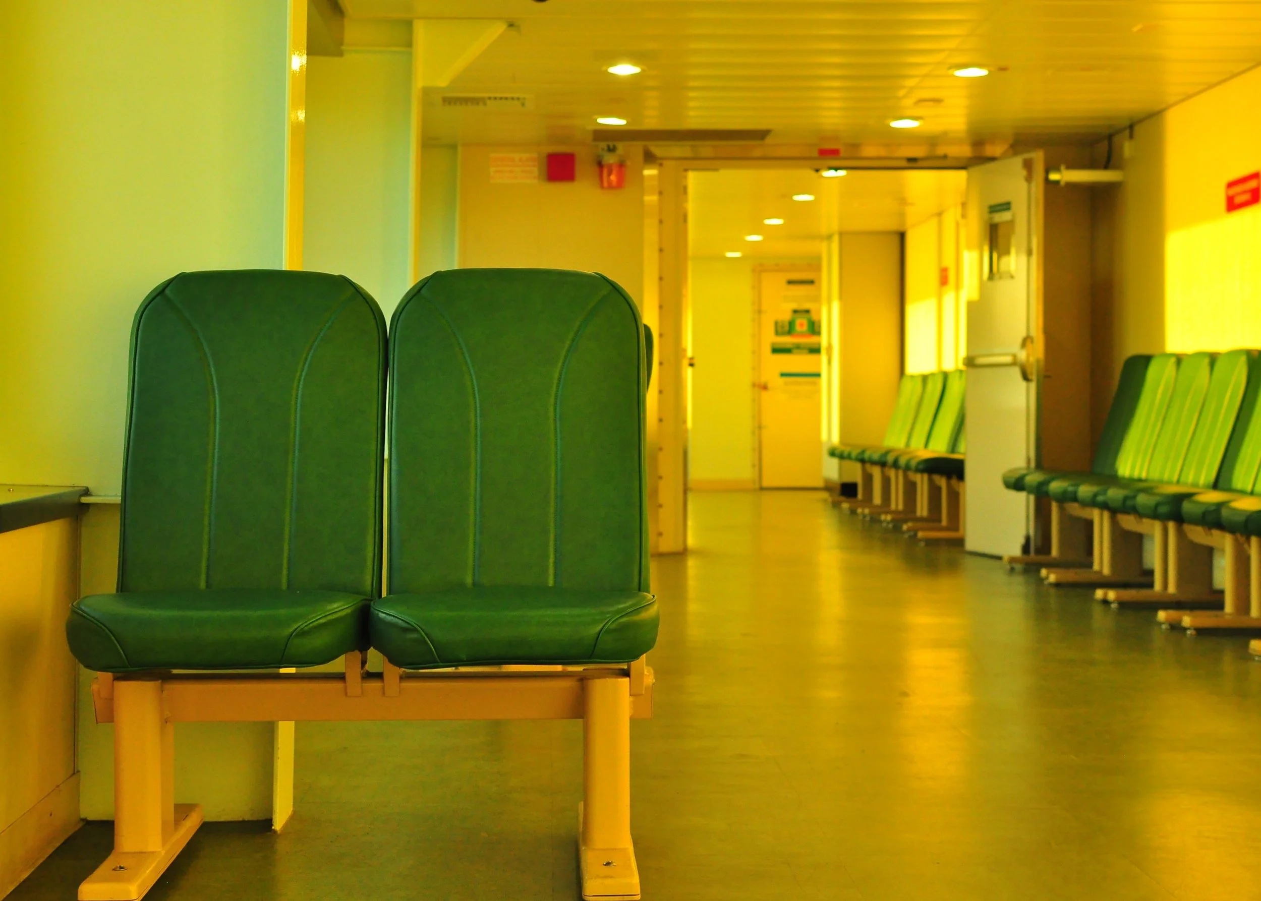Empty green bench seats in a waiting area of a ferry with additional green seats along the wall and overhead lights illuminating the space.