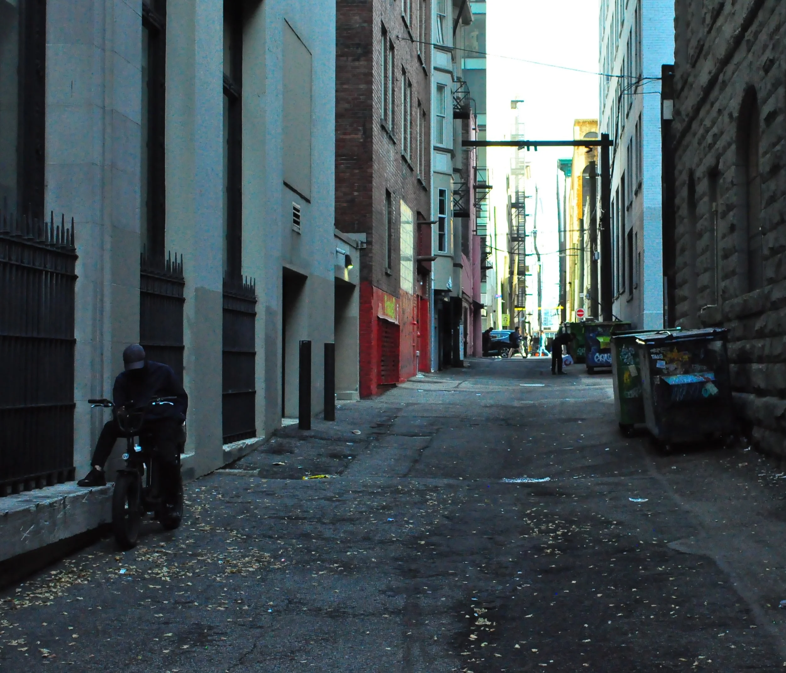 An alleyway between tall city buildings In Vancouver, BC with a person sitting on a small motorcycle on the left, and trash bins on the right, in downtown