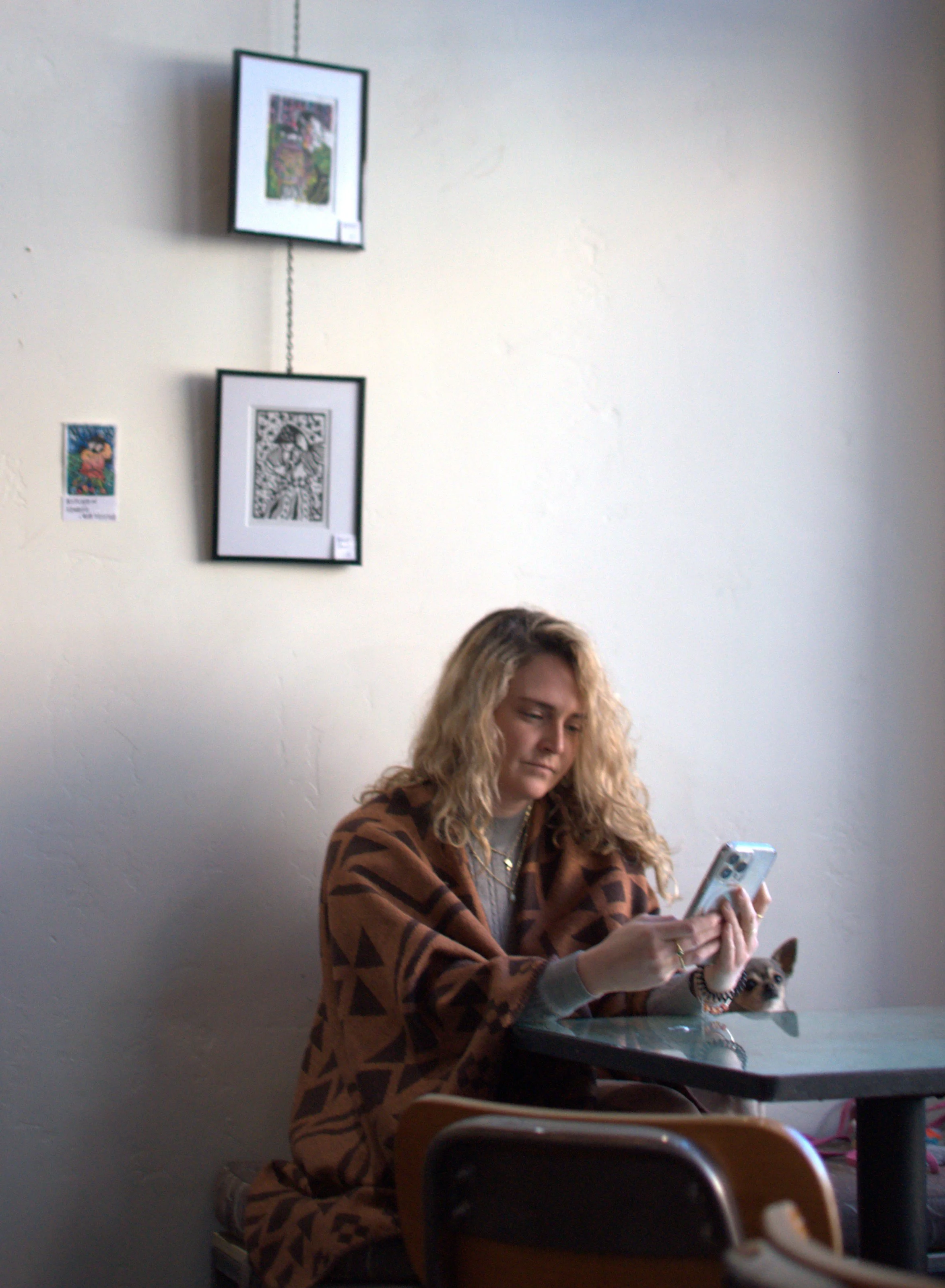 A woman with curly blonde hair wearing a patterned brown and black shawl sits at a table, looking at her phone. A small dog peeks out from behind her, resting on the table.