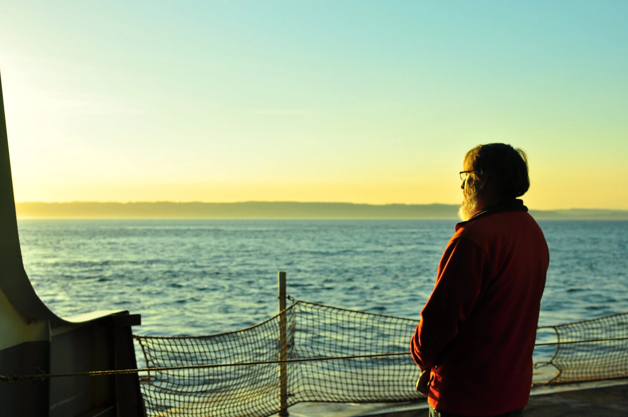 A man with glasses and a beard wearing a red jacket, standing on a boat or ferry, gazing at the lake or sea during sunset.