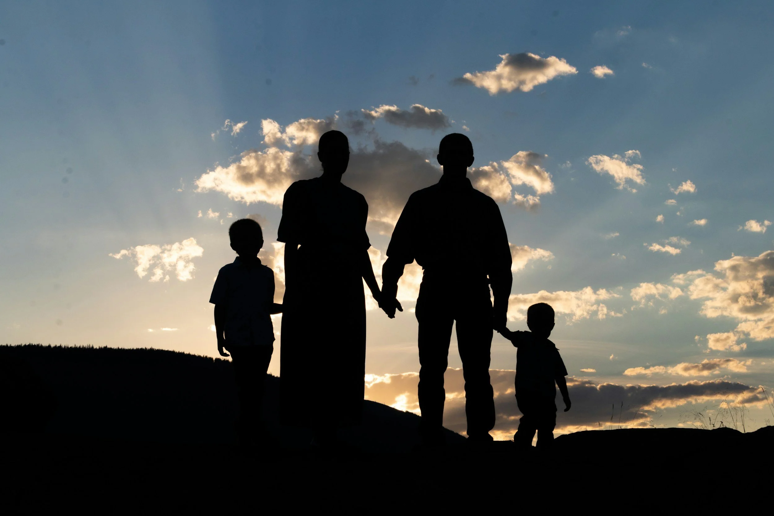 Silhouettes of a family holding hands walking outdoors during sunset with a sky filled with clouds.