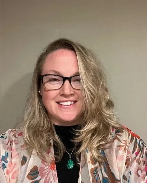 A woman with glasses and wavy blonde hair smiling at the camera, wearing a floral-patterned jacket over a black top and a green pendant necklace, standing against a plain light-colored wall.
