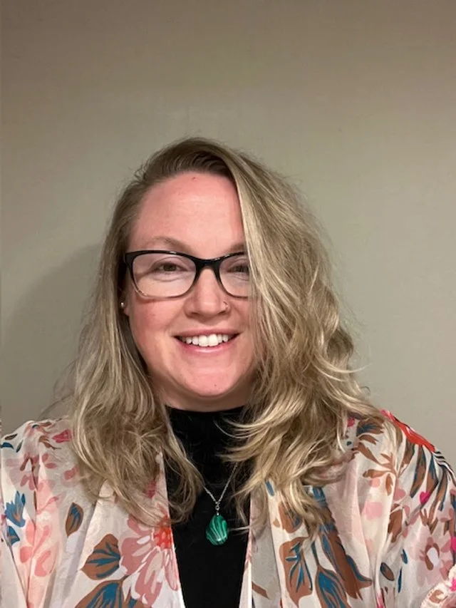 A woman with blonde, curly hair and glasses smiling at the camera, wearing a floral patterned blazer and a green pendant necklace against a plain wall background.