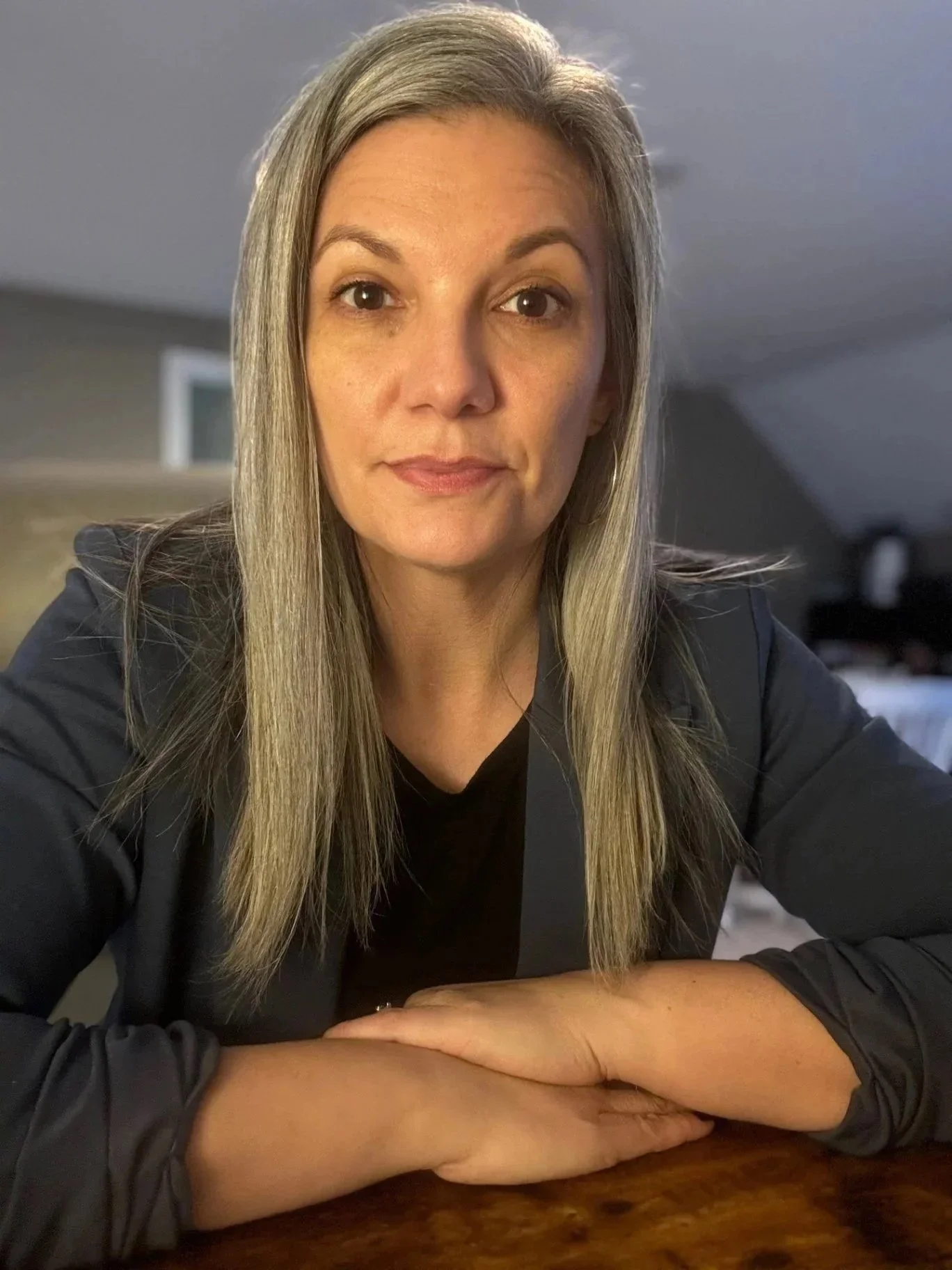 A woman with long blonde hair and fair skin looking into the camera, sitting at a wooden table with her arms crossed, in a room with gray walls and a window in the background.