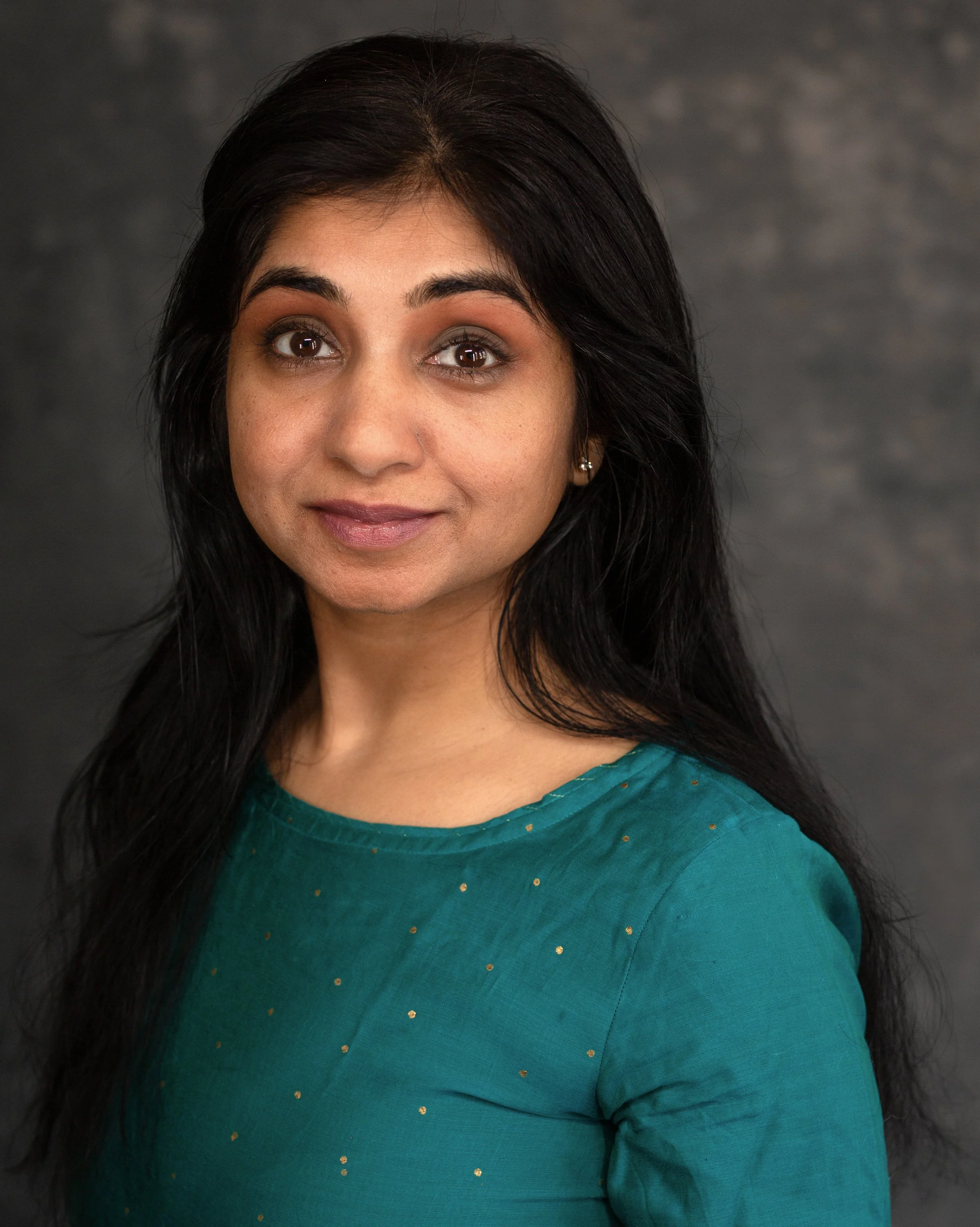Headshot of a woman with dark hair wearing a teal top with small golden dots, against a dark background.