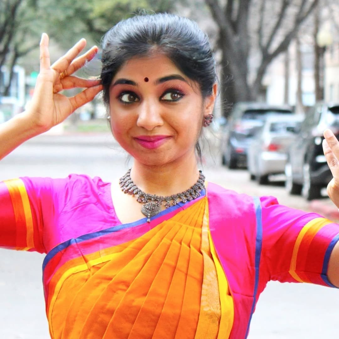 A woman with dark hair in a bun, wearing a colorful traditional sari and jewelry, making a playful gesture outdoors with parked cars and trees in the background.