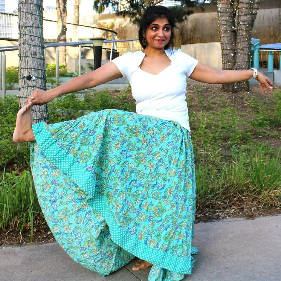 A woman doing a yoga pose outdoors, balancing on one foot while holding her other foot behind her with her hand. She is smiling and dressed in a white top and a long, blue floral skirt. There are trees and string lights in the background.