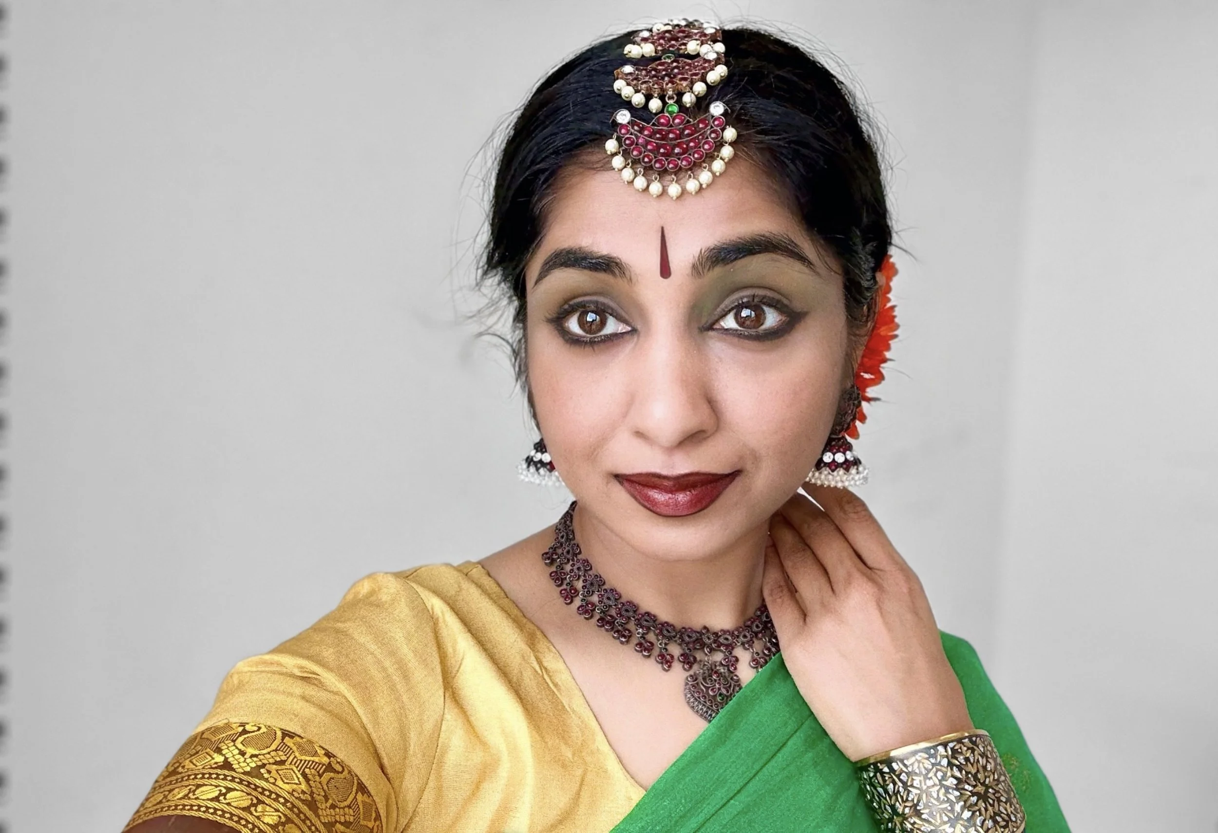 A woman dressed in traditional Indian attire, wearing a yellow and green sari with jewelry including a headpiece, earrings, necklace, bangles, and a red bindi on her forehead.