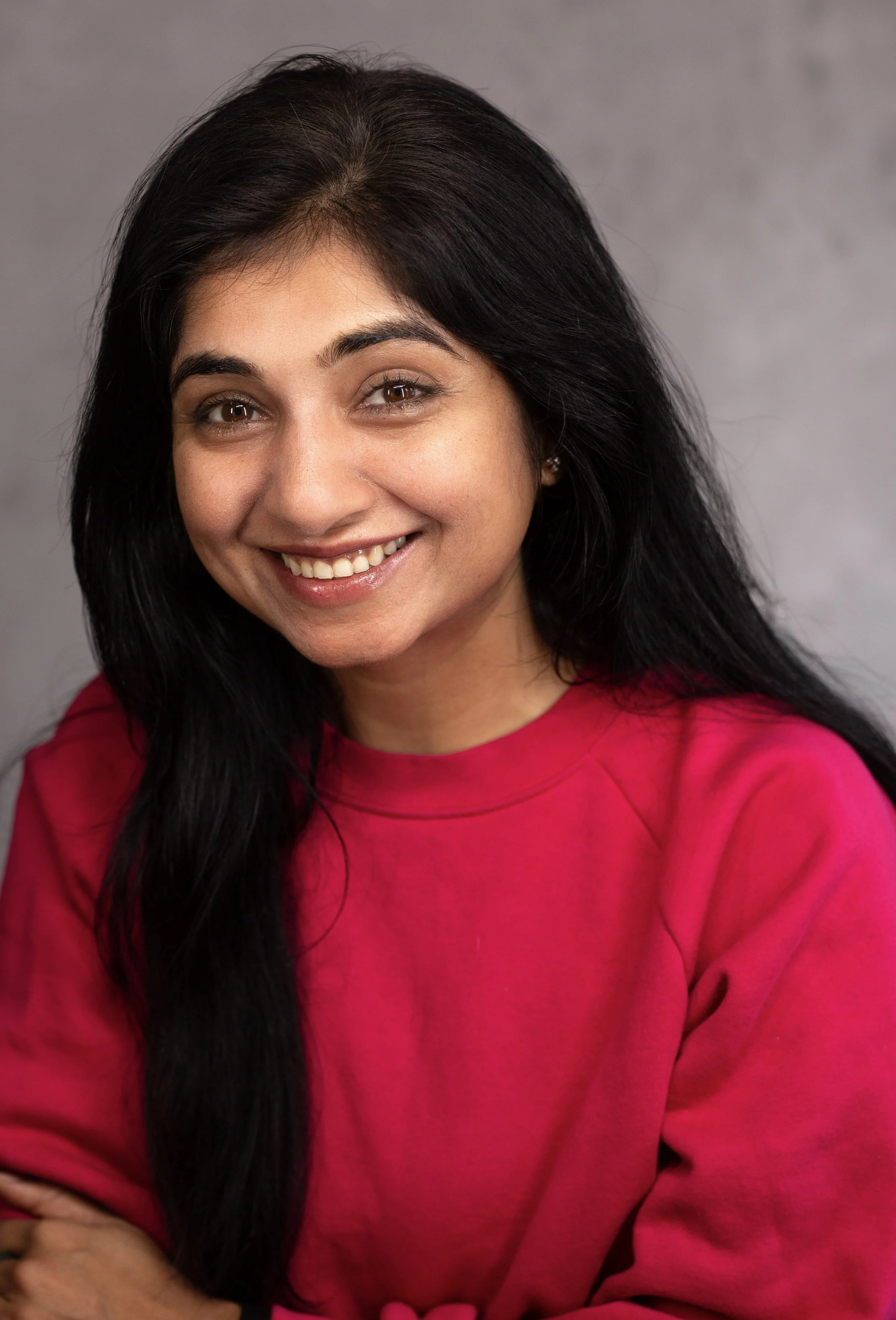 A woman with long black hair smiling, wearing a red sweatshirt, against a gray background.