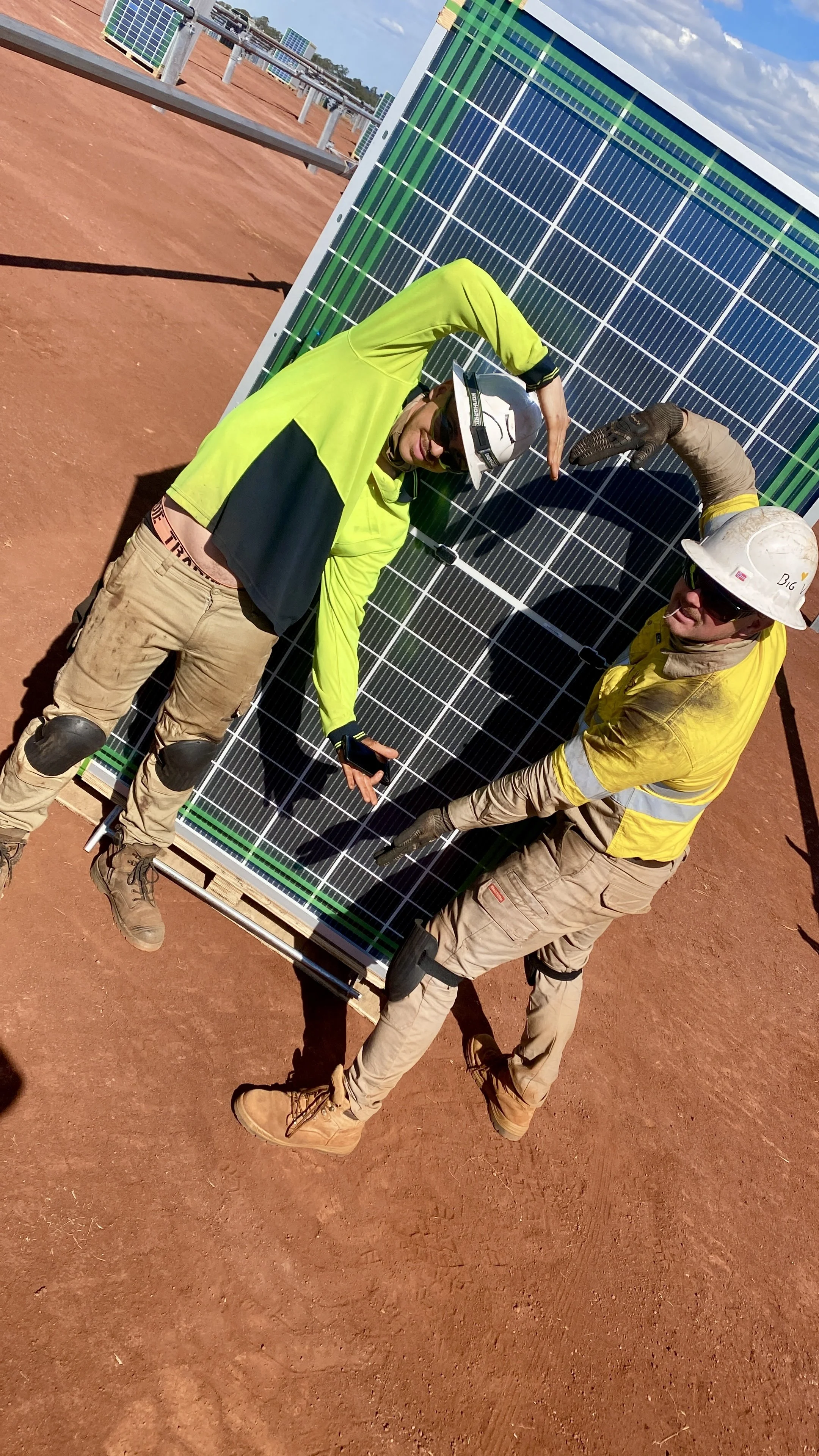 Two construction workers posing next to a large solar panel on a reddish dirt ground at a solar farm.