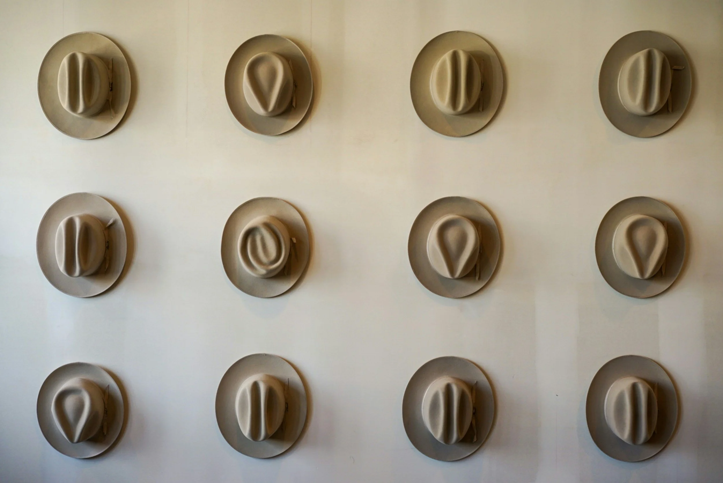 Multiple beige hats with different hatbands displayed on a wall in a grid pattern.