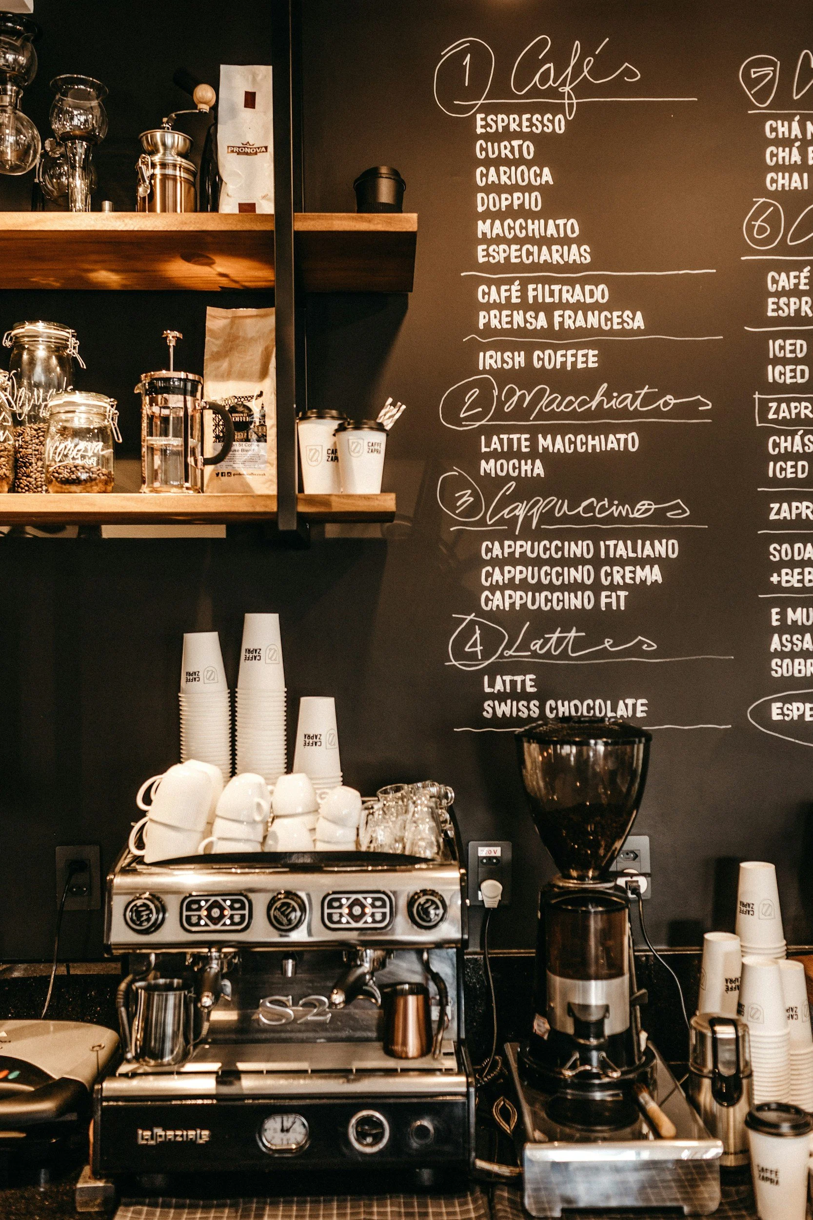 Coffee shop counter with an espresso machine, coffee cups, a coffee grinder, and a chalkboard menu listing various coffee beverages.