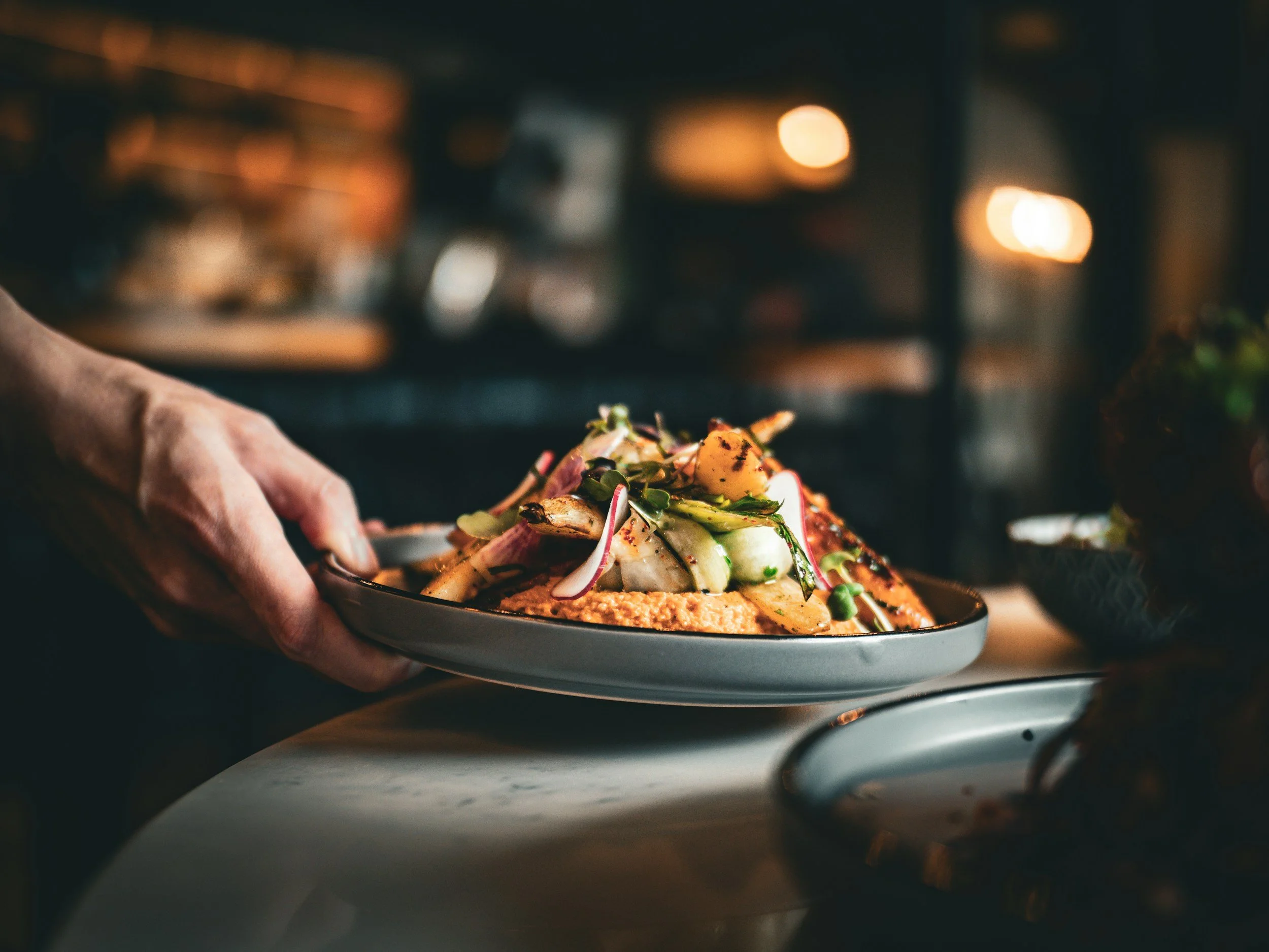 Person placing a plate of grilled vegetables and hummus on a table in a dimly lit restaurant setting.