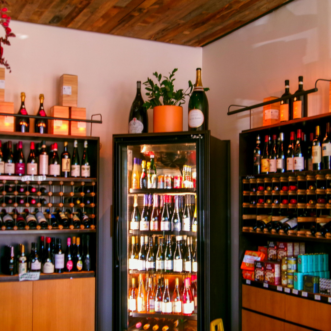 A wine shop with shelves of wine bottles, a wine cooler in the center filled with various bottles, and decorative bottles on top of the shelves. There's a potted plant and wooden boxes on top of the shelf.