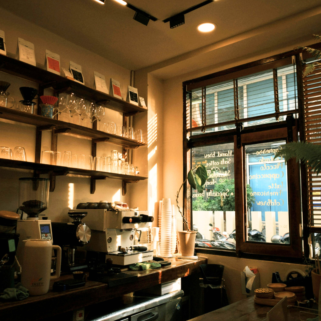 Interior of a cozy coffee shop with shelves of glassware, espresso machine, and a window with blinds letting in natural light.