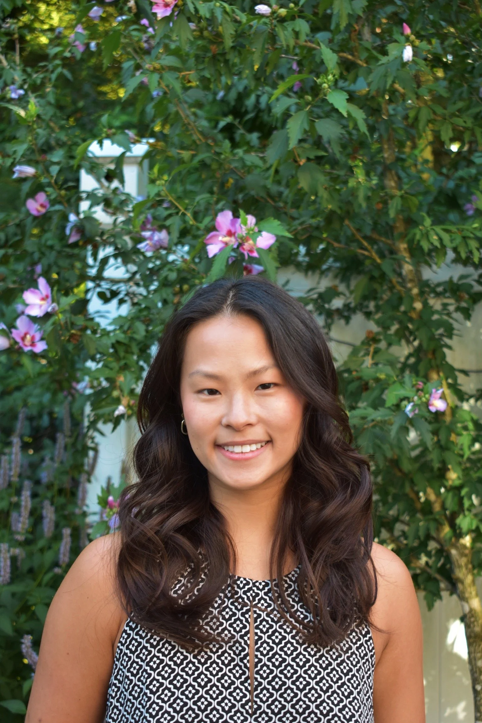A young woman with long, dark, wavy hair smiling in front of a bush with pink flowers.