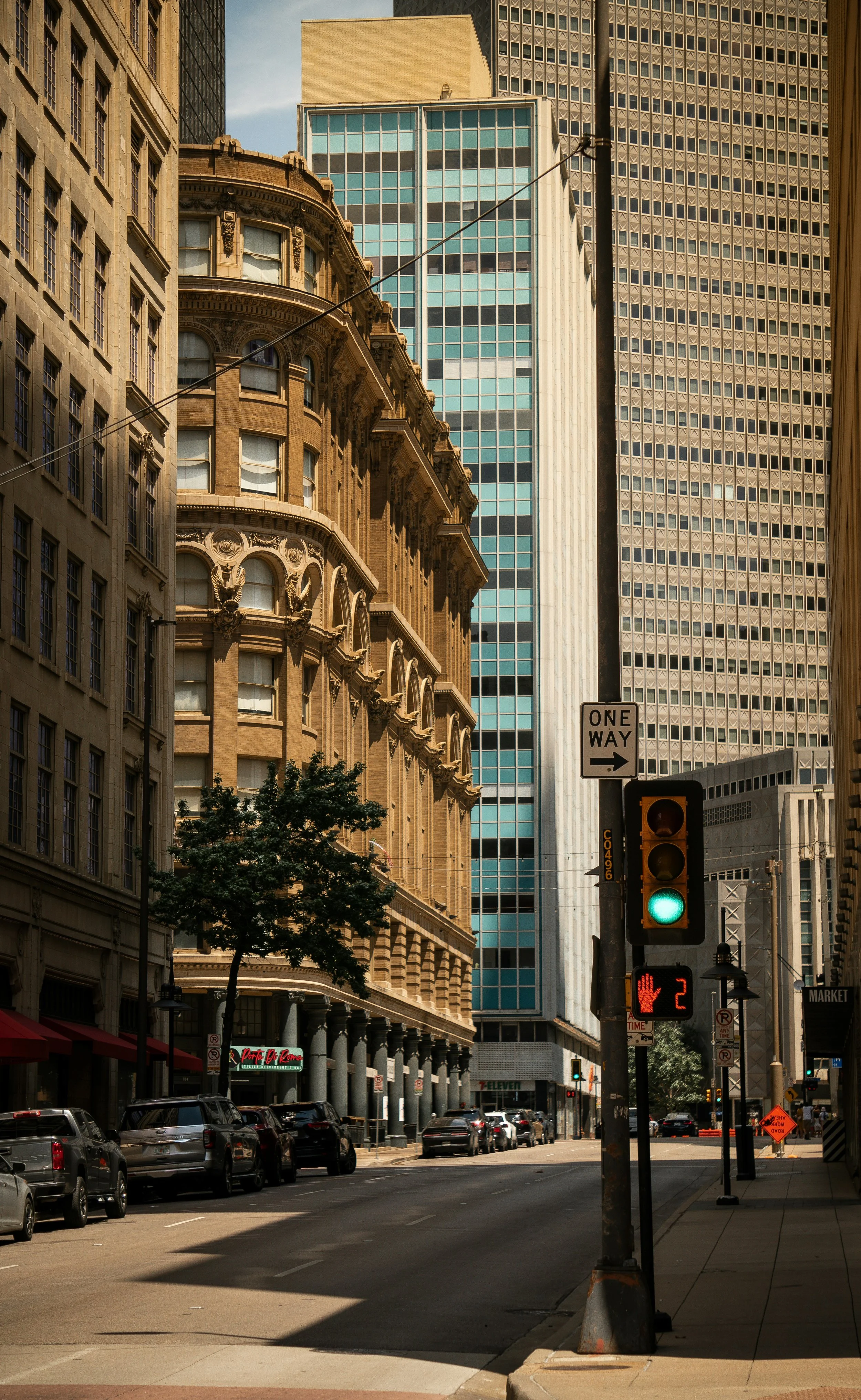 City street with tall buildings, parked cars, traffic light showing green, pedestrian signal with a hand and the number 2, and a tree in the foreground.