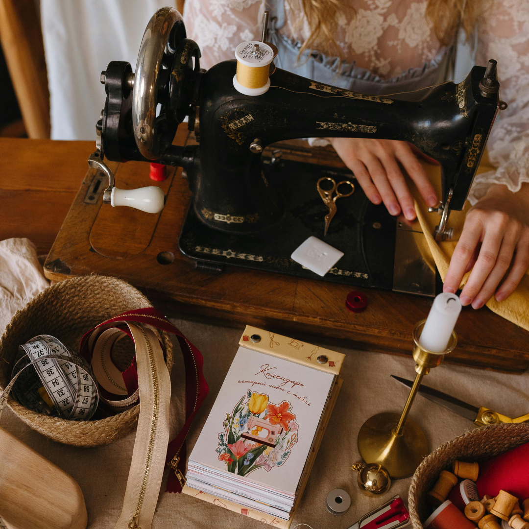 Person sewing with a vintage black sewing machine on a wooden table, surrounded by sewing supplies like measuring tape, ribbons, scissors, and thread.