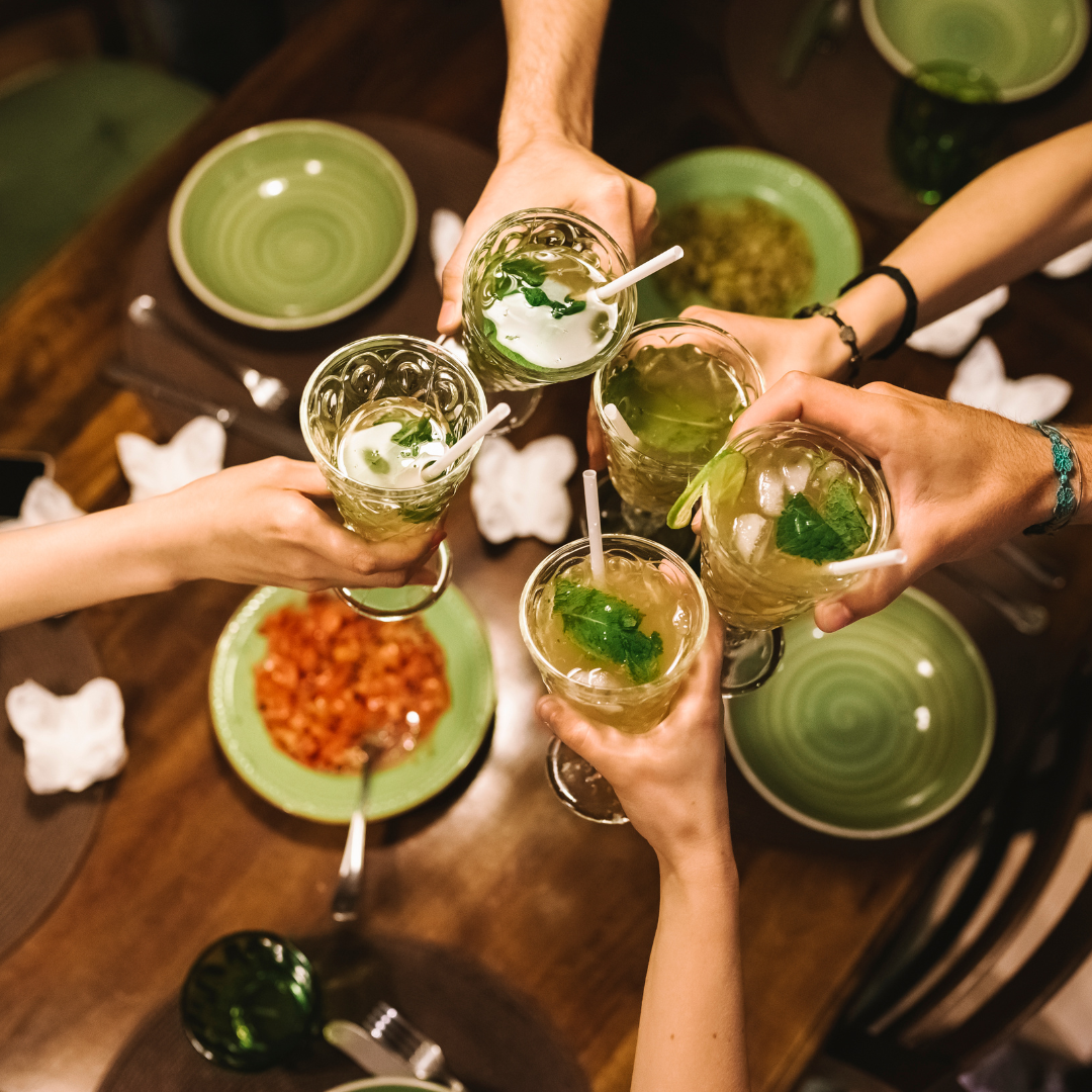 Six people raising glasses of mint lemonade with ice and straws in a toast, on a wooden table with green and white plates and a dish of salsa.
