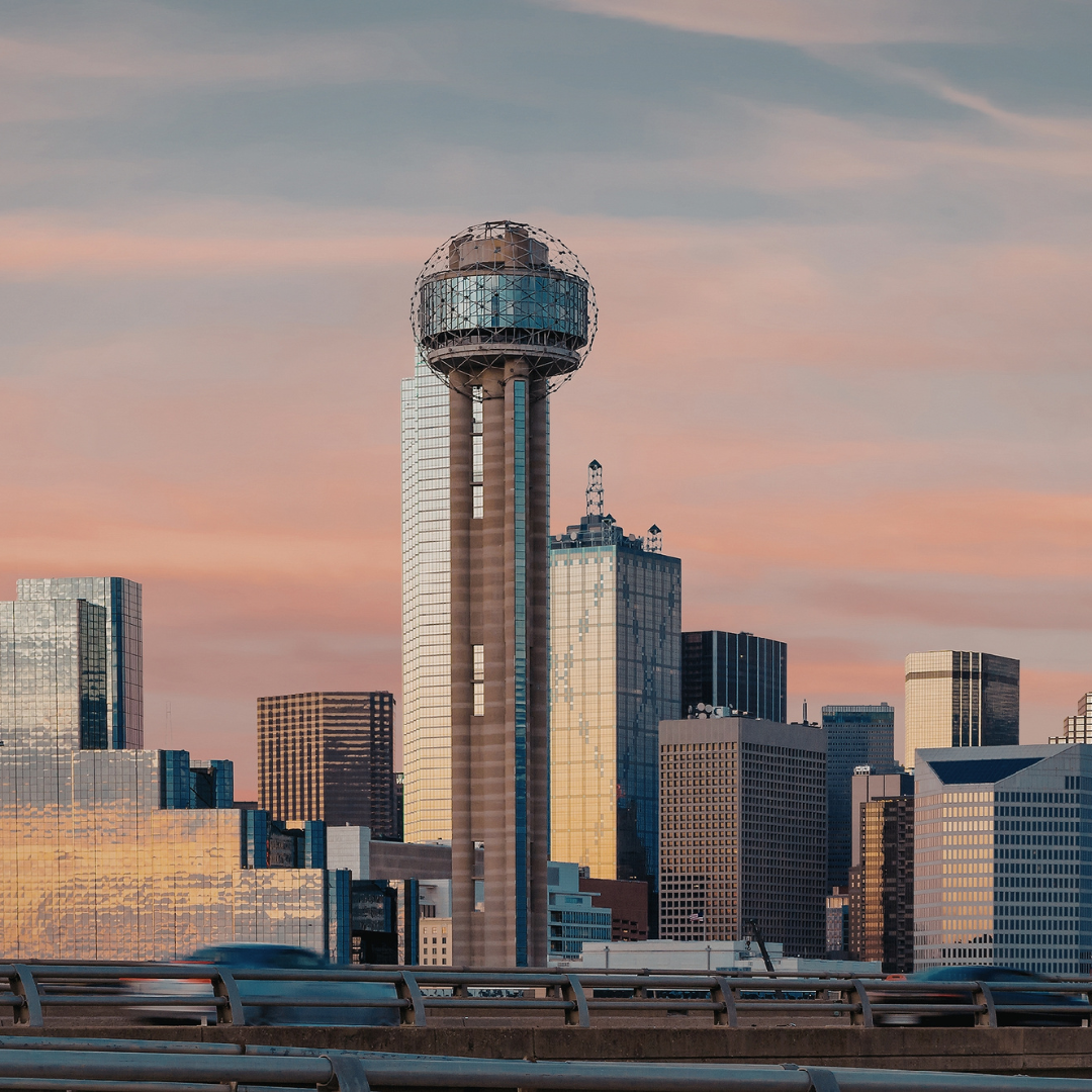 City skyline with a tall tower featuring an observatory or spherical structure at the top, during sunset with pink and blue sky.