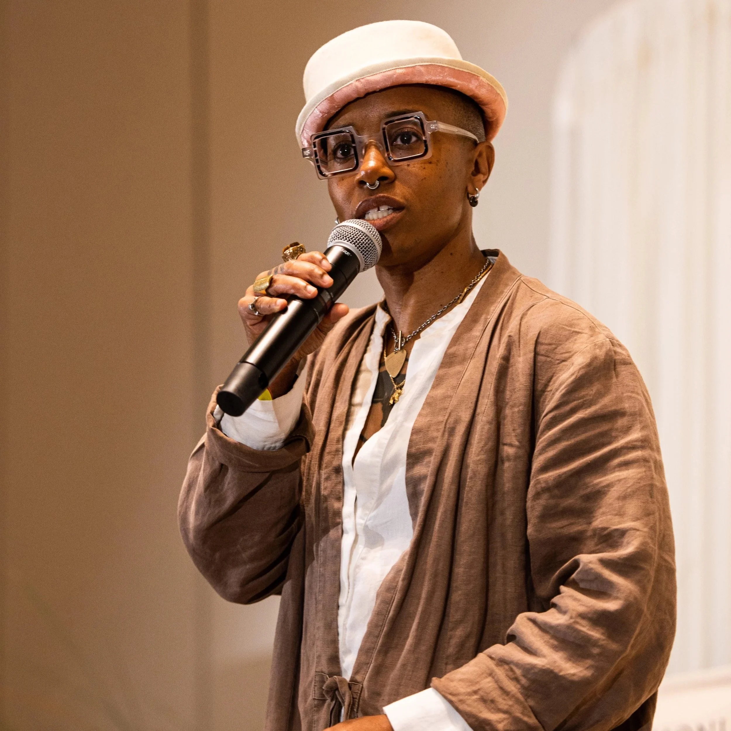 A woman wearing glasses, a pink hat, and various jewelry, speaking into a microphone during an event.