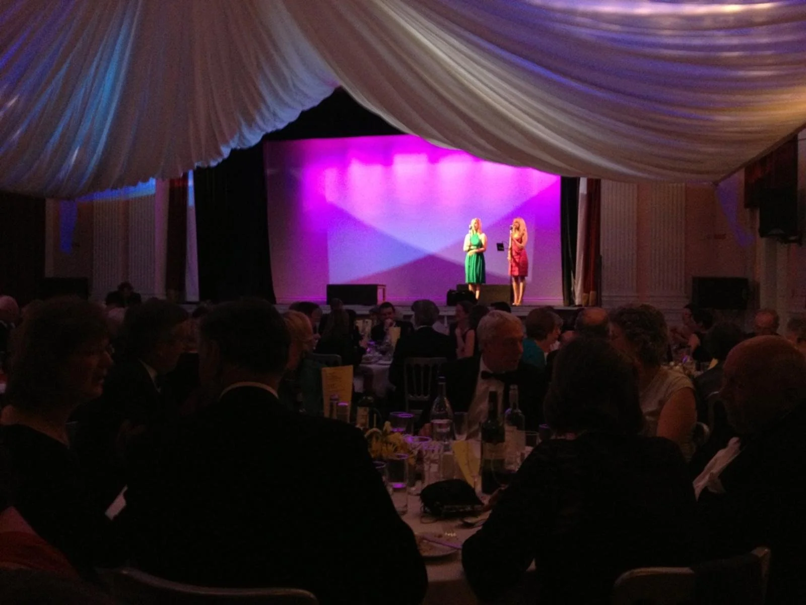 Two women stand on stage in front of an audience seated at round tables, with a pink and purple backdrop, under a large white canopy. The women are engaged in a conversation or performance.
