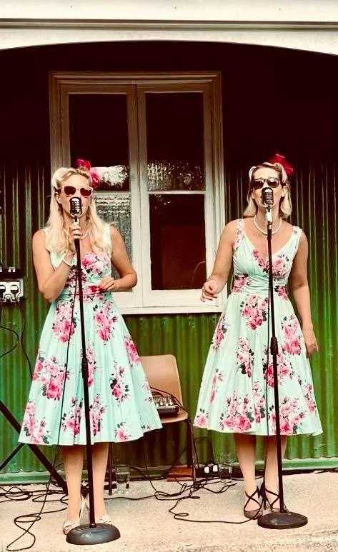 Two women wearing matching floral dresses, sunglasses, and red bows, singing into microphones during a performance.