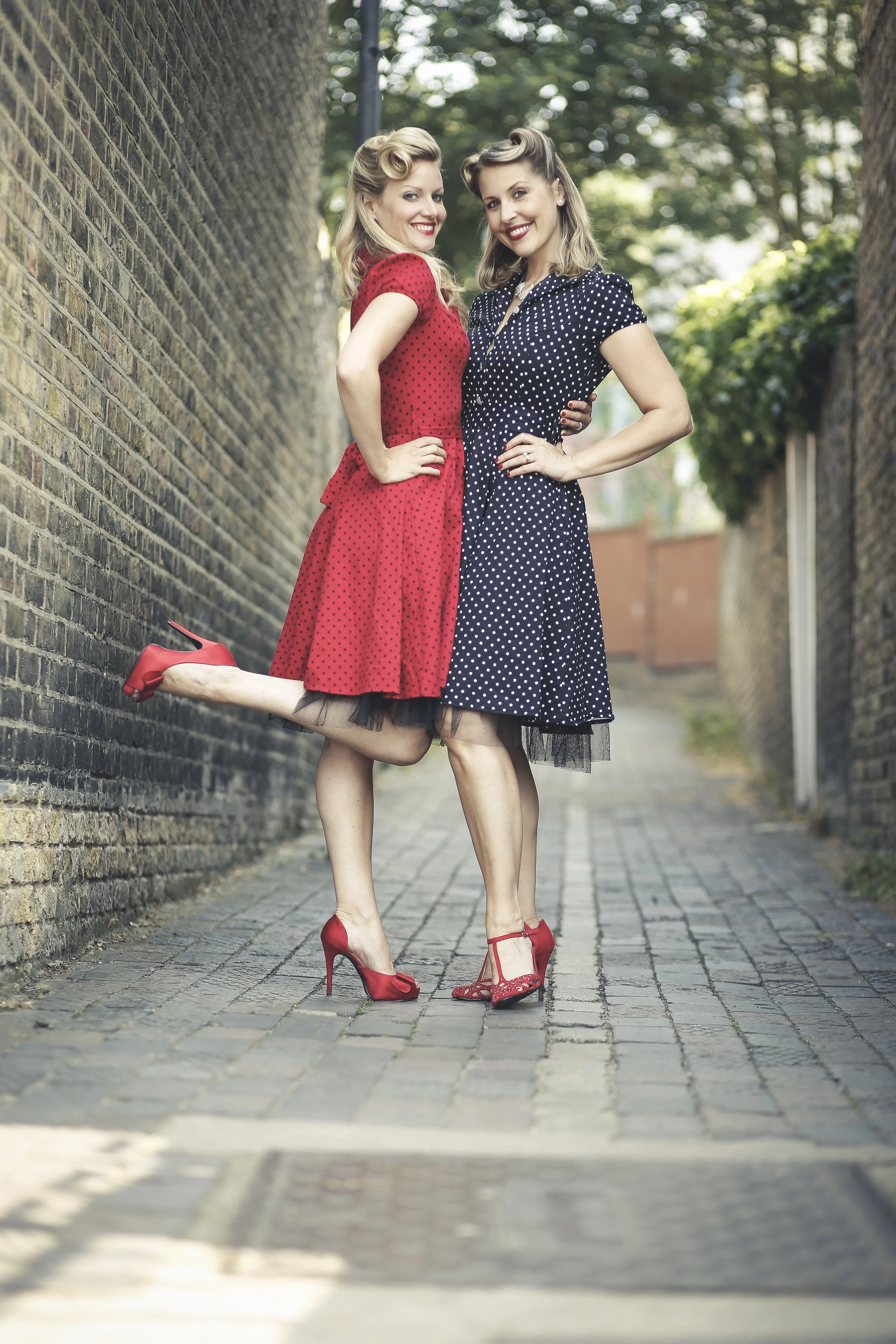 Two women in vintage dresses with polka dots, wearing red high heels, standing cheerfully on a cobblestone alleyway between brick walls, with trees in the background.