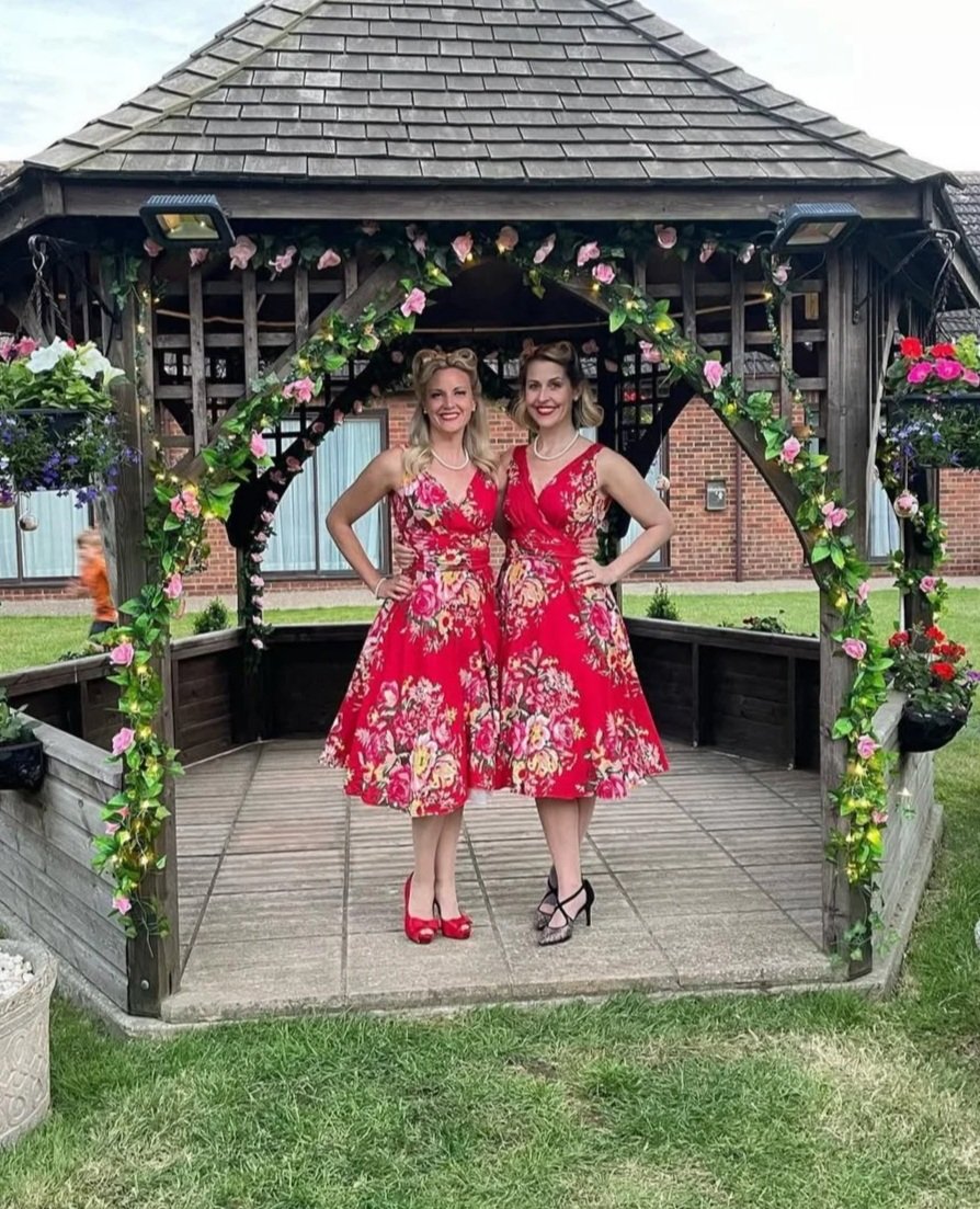 Two women in red floral dresses standing in a decorated gazebo, smiling at the camera.