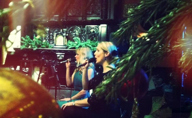 Two women singing into microphones on stage, surrounded by festive decorations and greenery, in a dimly lit setting.