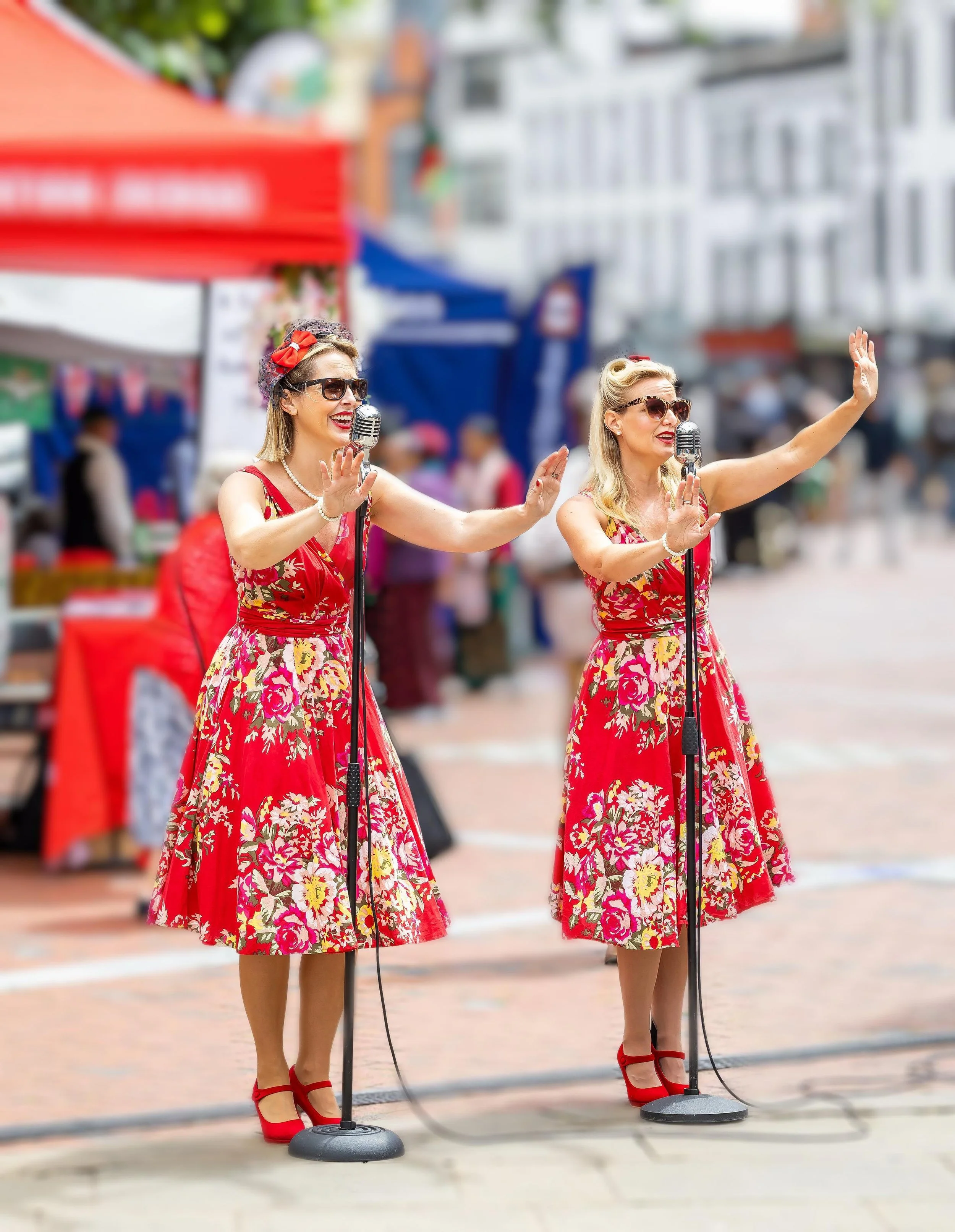 Two women in vintage red floral dresses and red heels singing into microphones at an outdoor event.