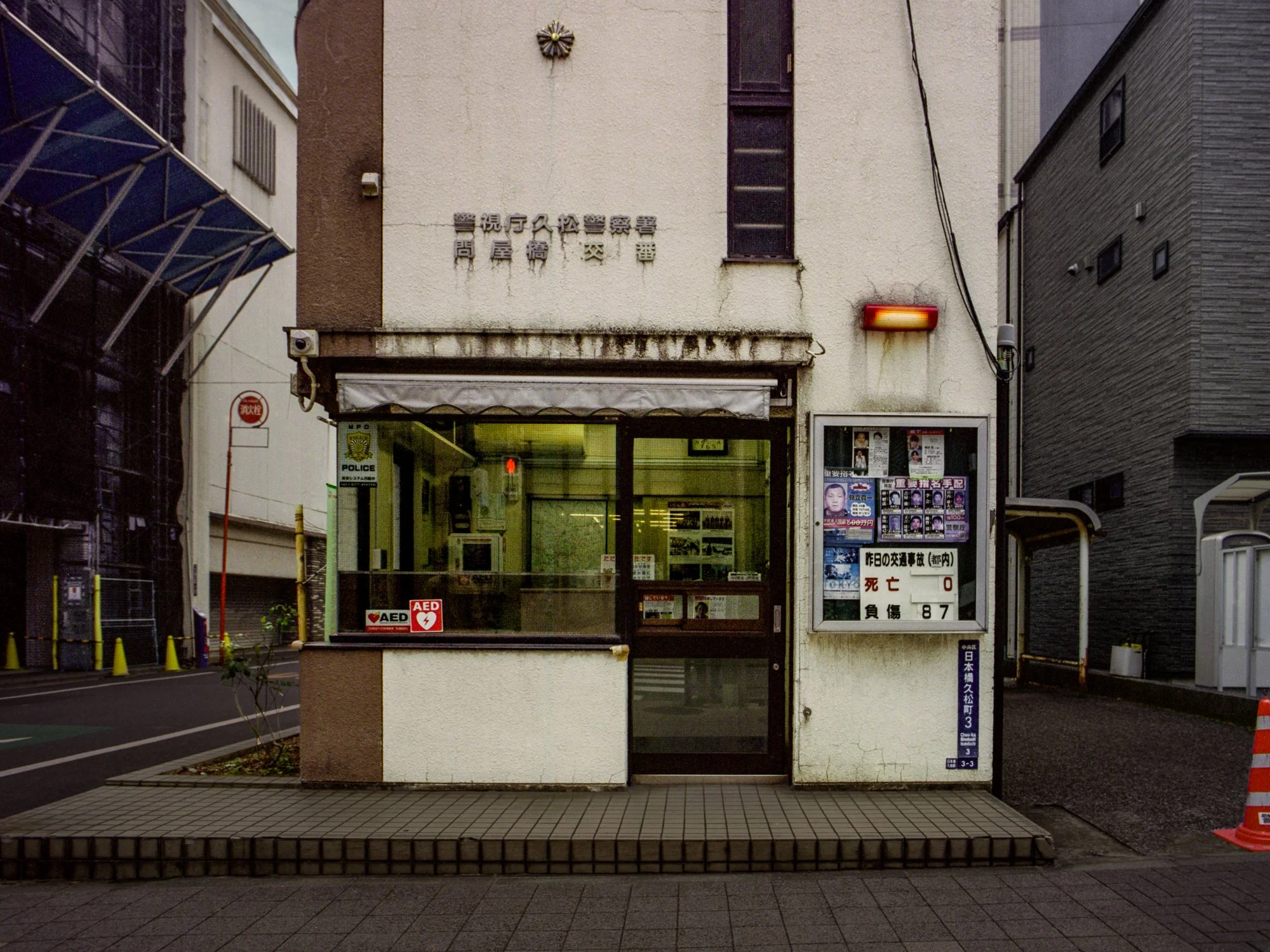 A small, Japanese community police station with a glass window and door, displaying a police sign and AED sticker, with posters and notices on the side board, and a yellow and black pole outside on an urban street.