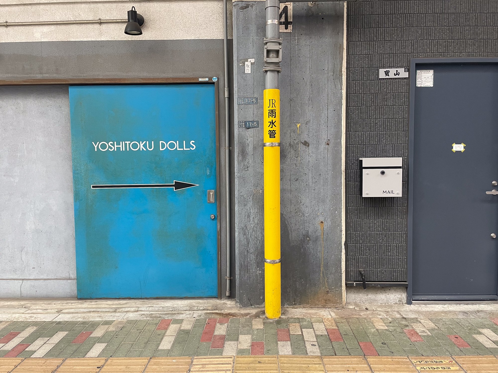 A blue door labeled 'YOSHITOKU DOLLS' with a black arrow pointing to the right. There is a yellow pole with Japanese characters, a mailbox labeled 'MAIL,' a gray door with a small sign, and a gray wall with a sign in Japanese. The ground is from a si