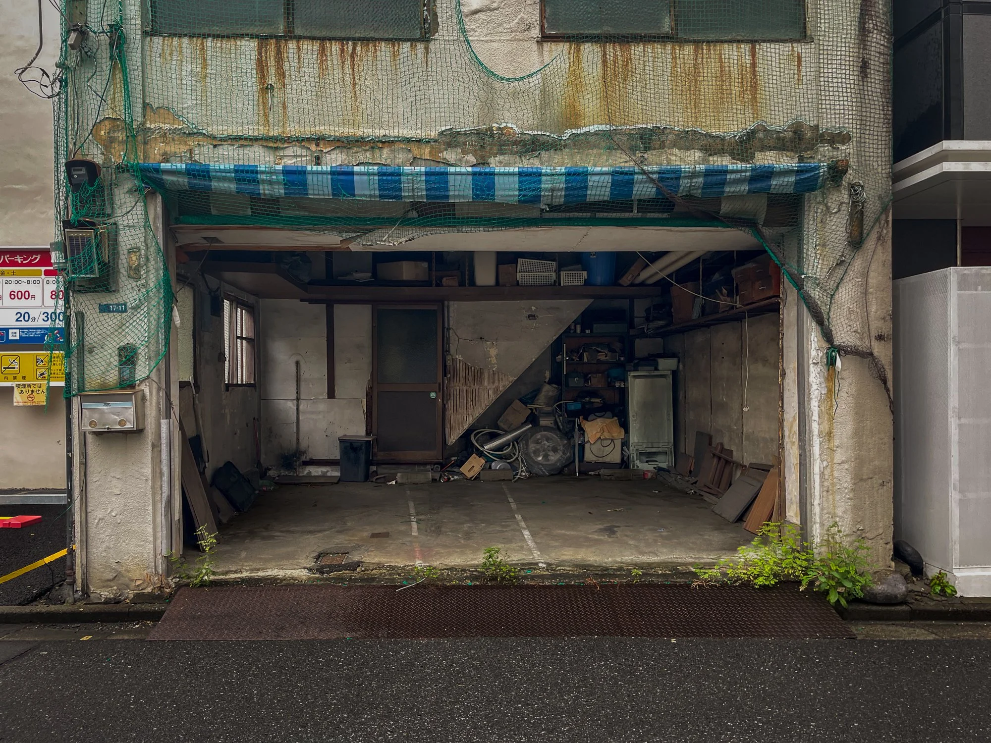 A small, empty garage with various clutter and items inside. The building has a rusted exterior with a striped awning and some greenery growing at the base. There are signs and a parking meter on the left side.