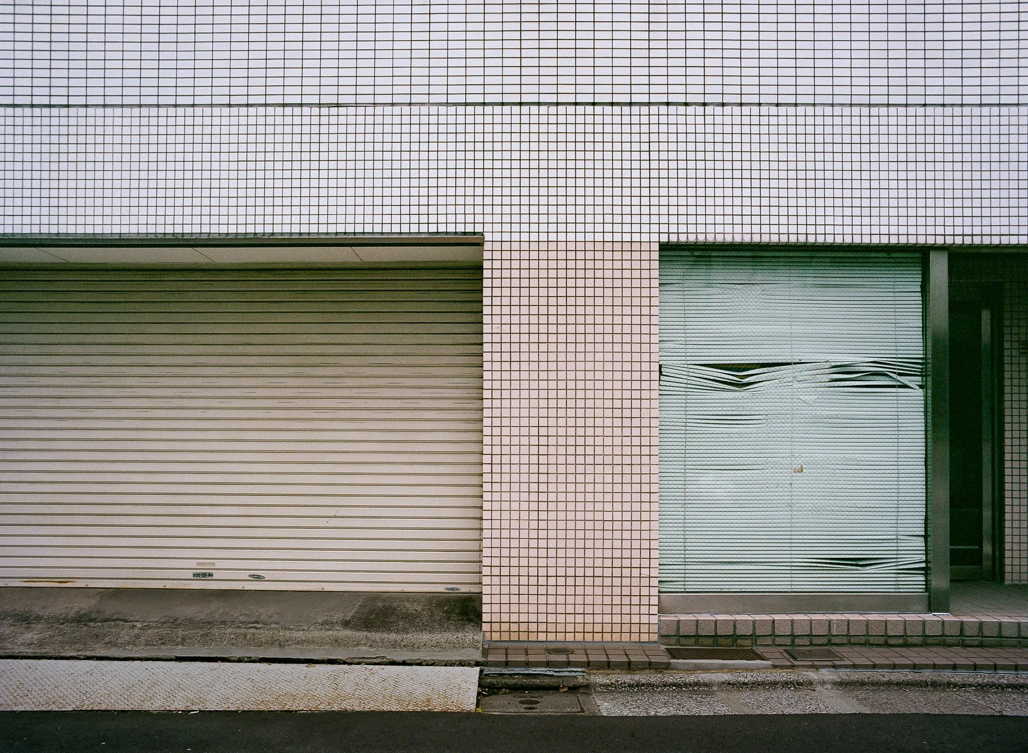 A building with a beige roller shutter door on the left and a glass window with a broken blind on the right. The exterior wall is covered in small pink tiles.