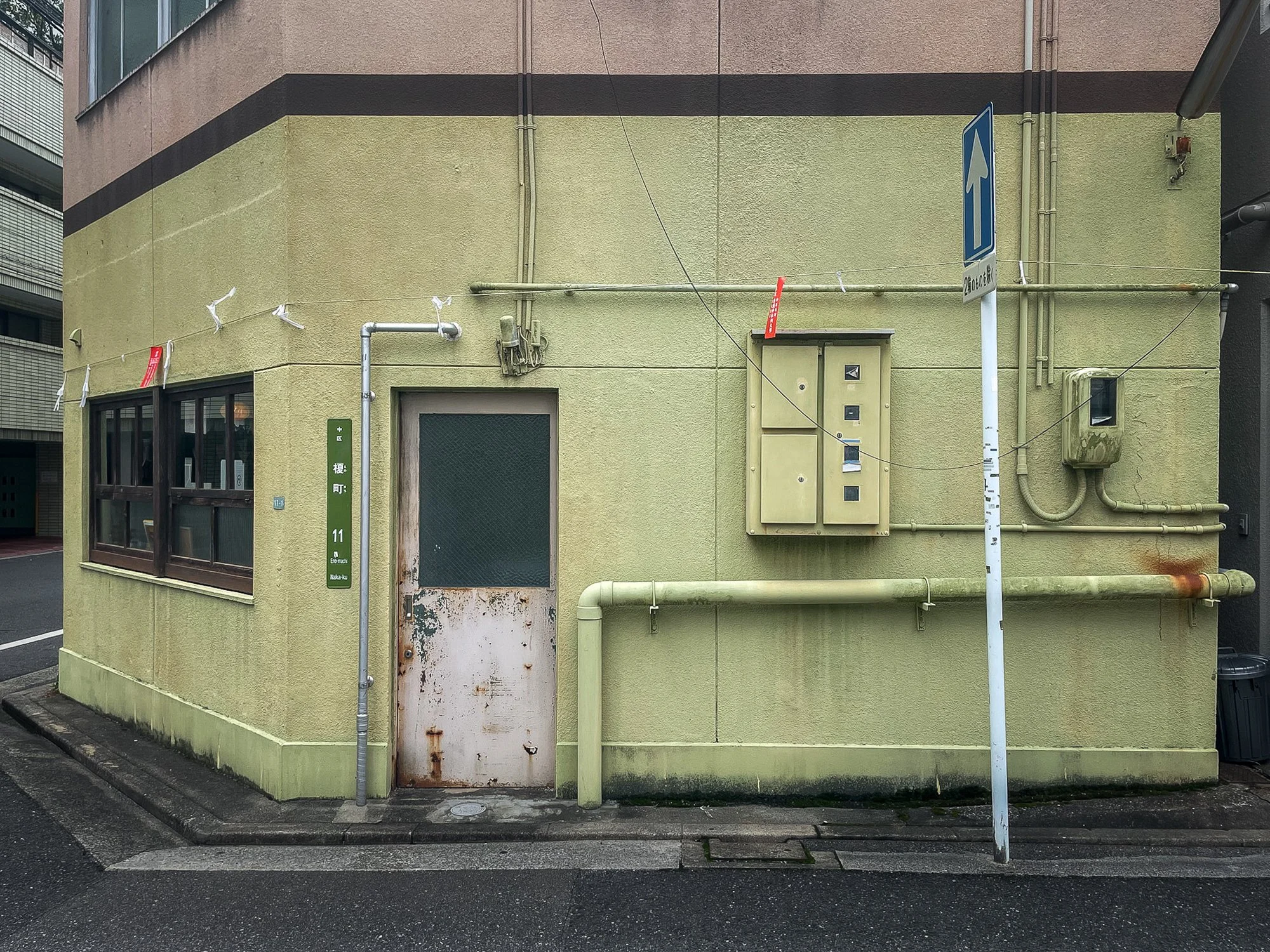 A small, green/yellow building with a rusty door, window, and various electrical boxes and pipes on the exterior wall. There is a green street sign and a blue one-way sign, with street and sidewalk visible in an urban setting.