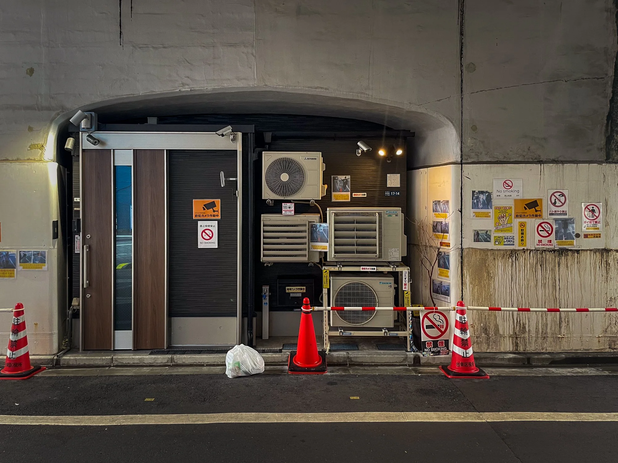 Underpass wall with air conditioning units, warning signs, and covered notices, with a no smoking sign and orange traffic cones, some caution tape, and a plastic bag on the ground.